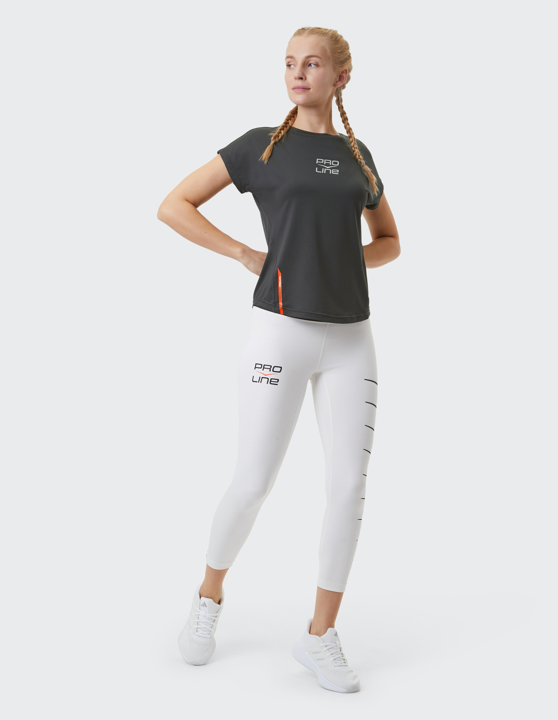 Frau in schwarzem Sportshirt und weißen Leggings mit "Pro Line"-Aufdruck, trägt weiße Sneakers, steht selbstbewusst mit verschränkten Armen.