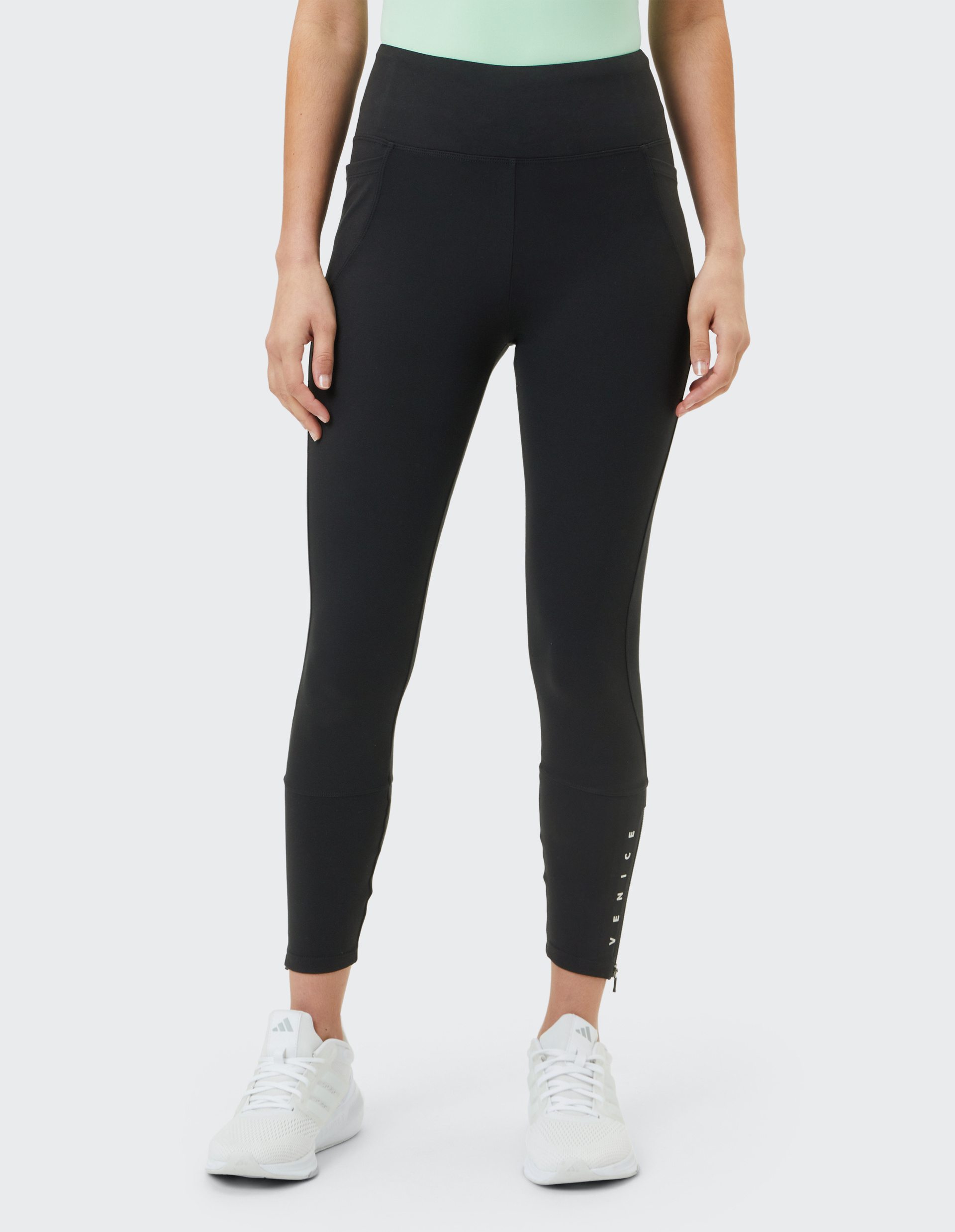 Person trägt schwarze Leggings mit hohem Bund und Reißverschlussdetails an den Knöcheln sowie weiße Turnschuhe auf grauem Hintergrund.