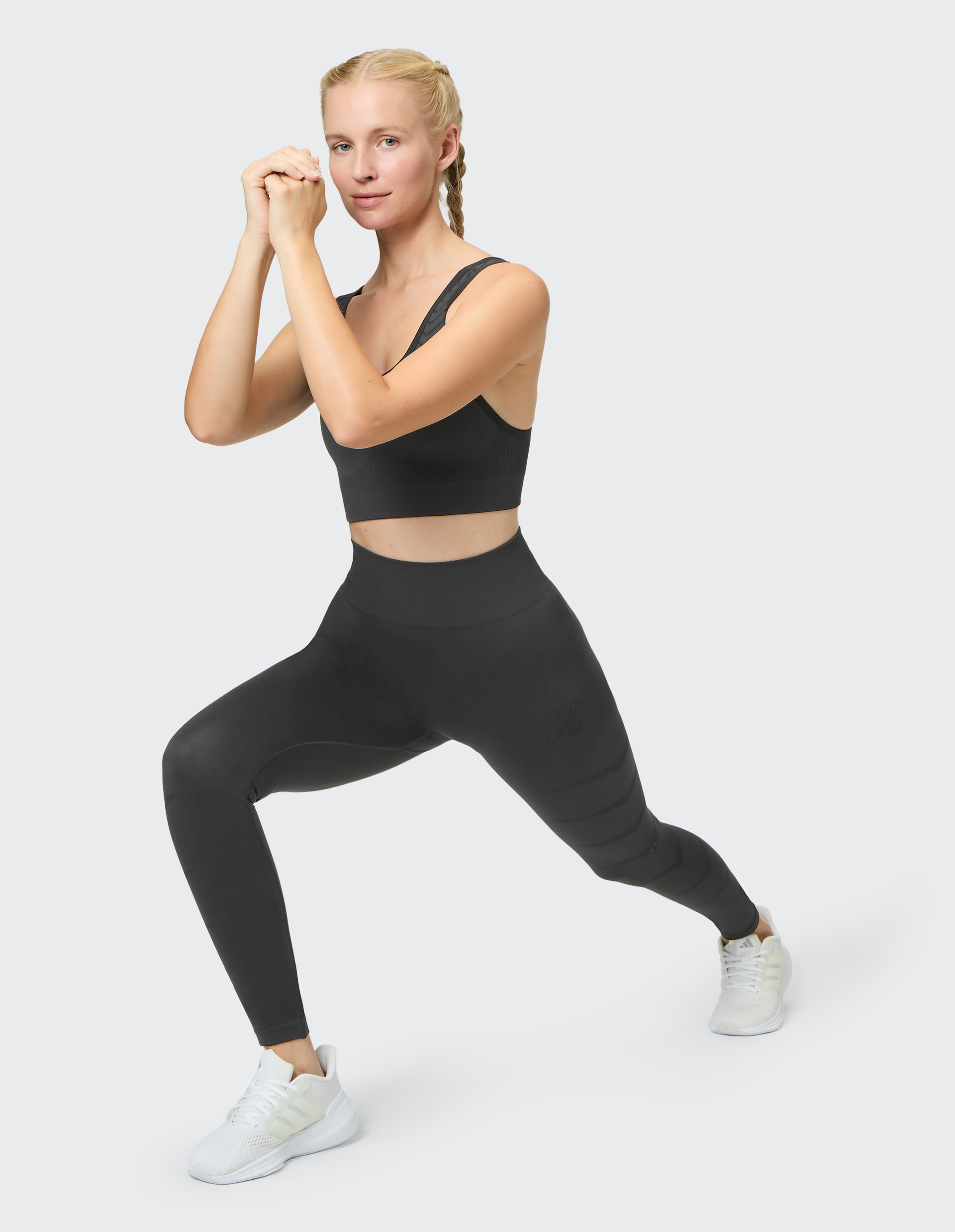 Frau in schwarzem Sport-BH und Leggings macht eine Ausfallschrittübung auf weißem Hintergrund, trägt weiße Turnschuhe.