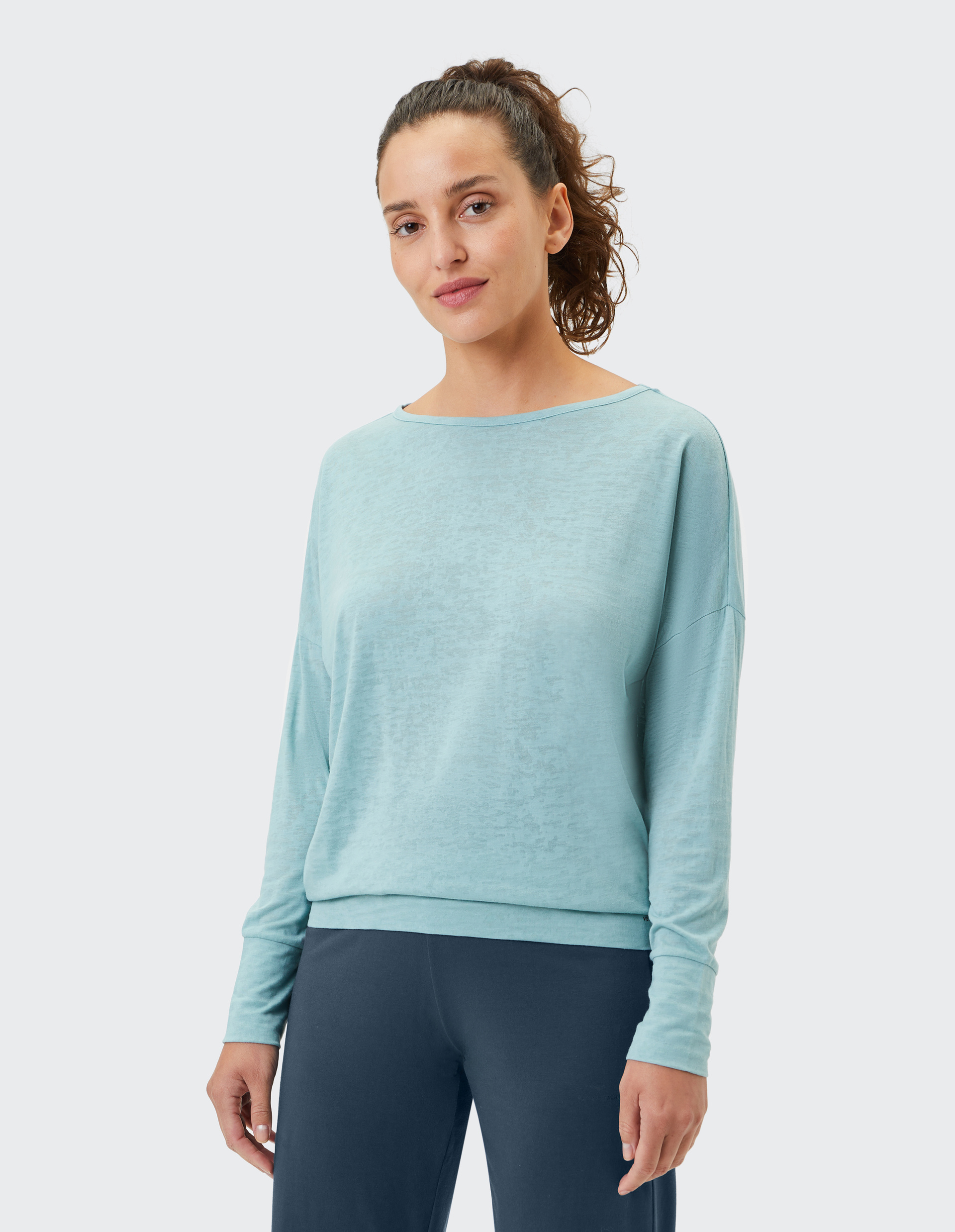 Eine Frau mit lockigem Haar trägt ein hellblaues Oberteil und dunkelblaue Hose vor einem neutralen Hintergrund.