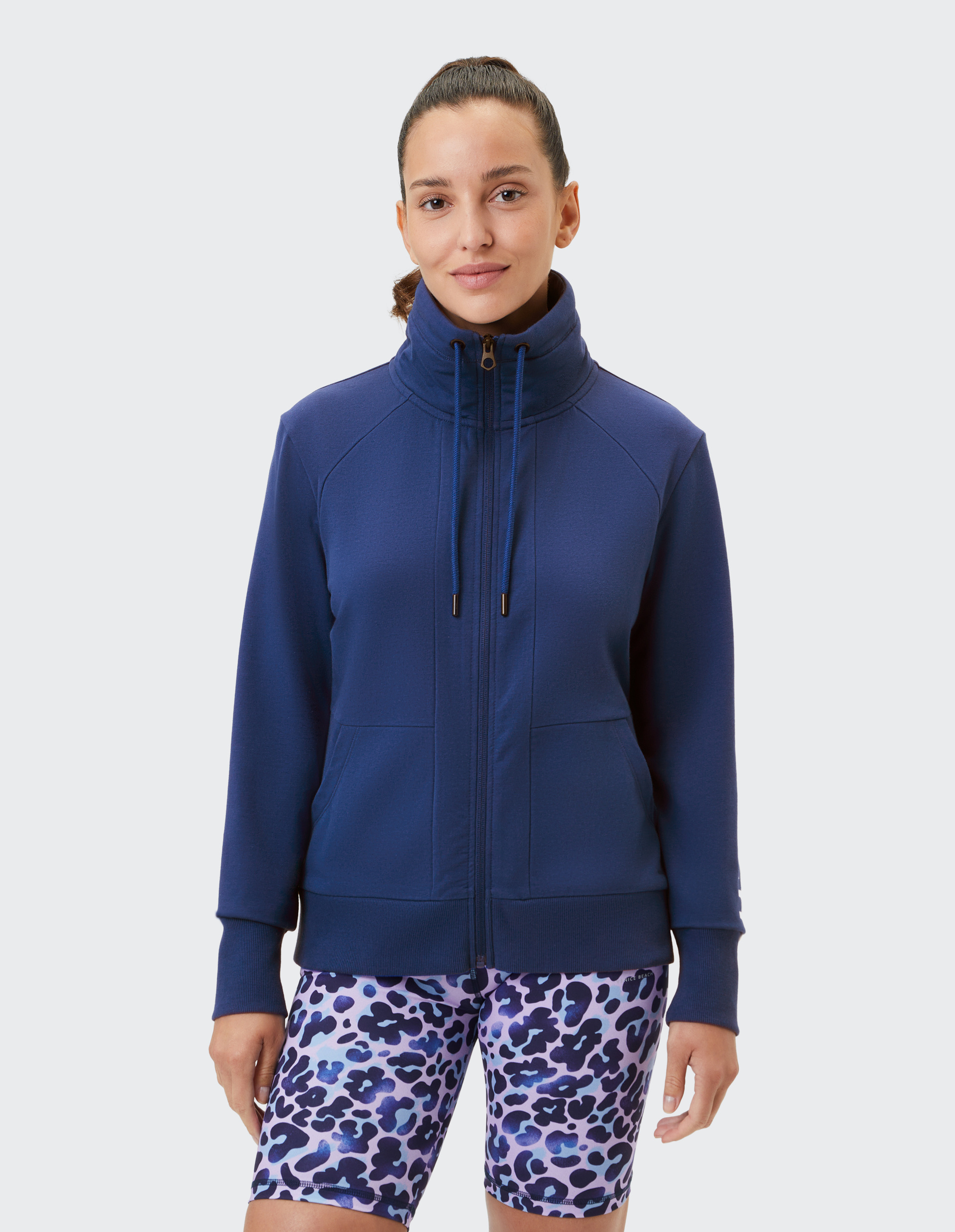 Frau in marineblauer Jacke mit hohem Kragen und lilafarben bedruckter Shorts, lächelt vor grauem Hintergrund in die Kamera.