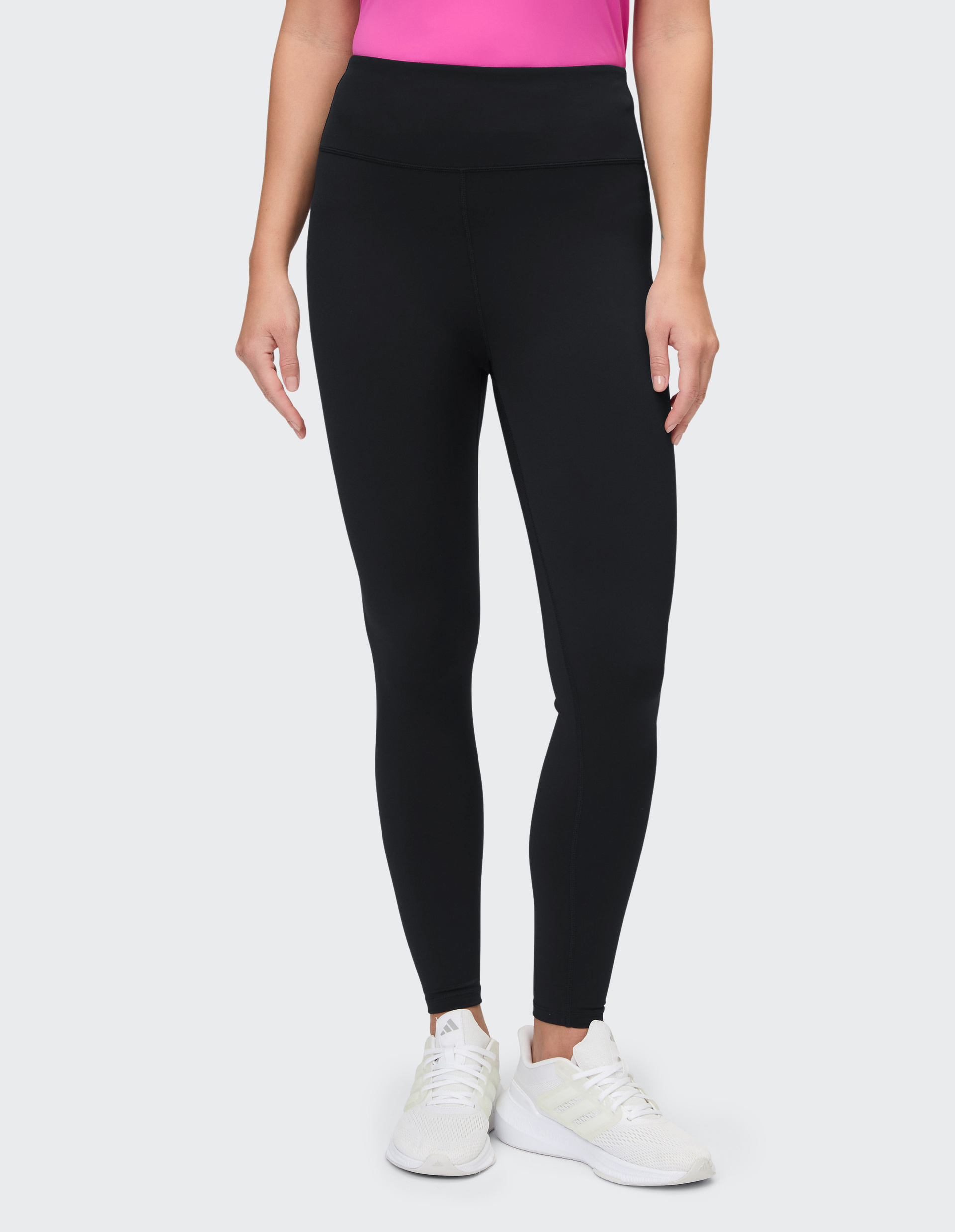 Eine Person trägt schwarze Leggings und weiße Turnschuhe, kombiniert mit einem pinken Oberteil, vor neutralem Hintergrund.