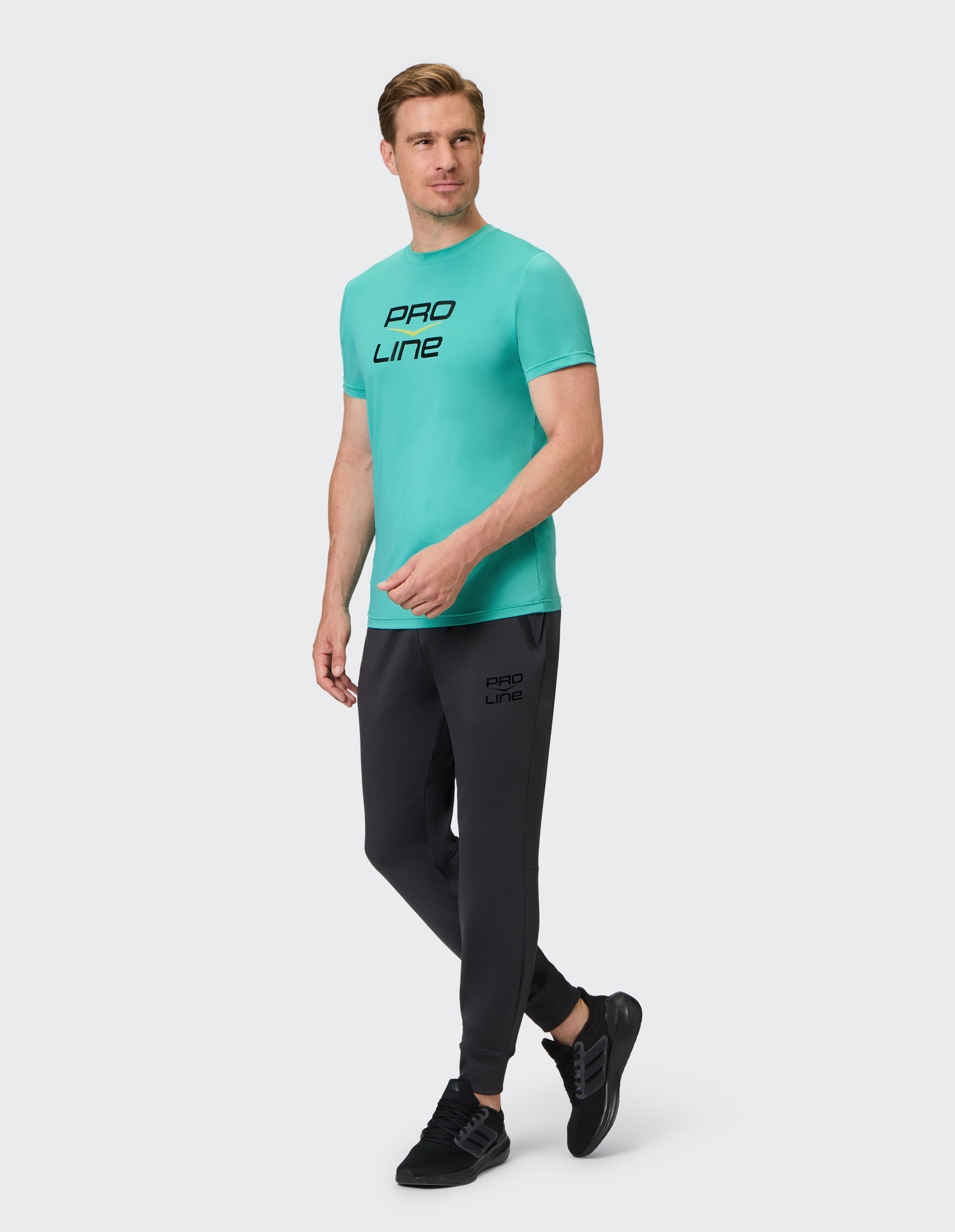 Mann in türkisfarbenem T-Shirt mit "PRO LINE"-Aufdruck und dunklen Jogginghosen, gehend vor hellem Hintergrund.