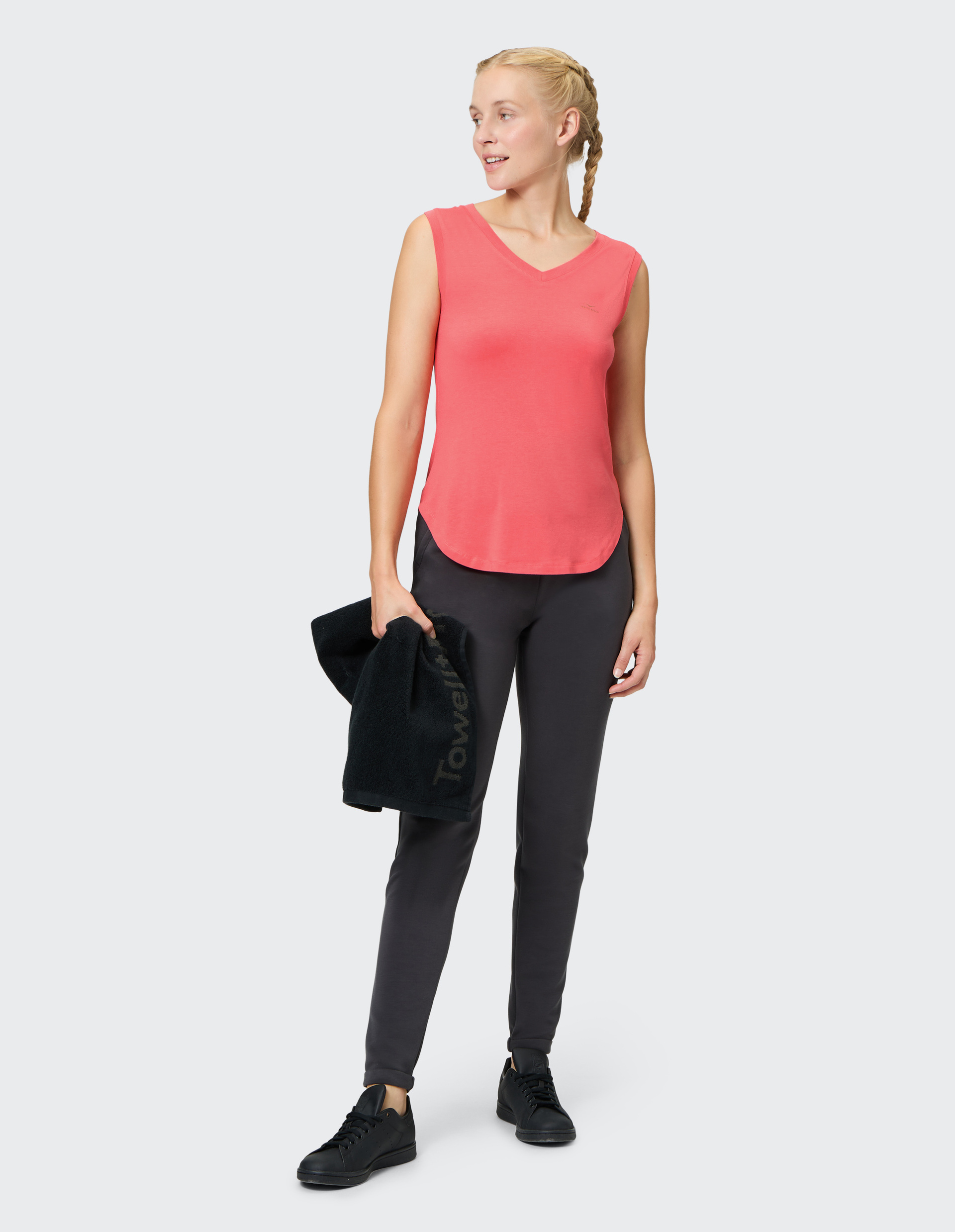 Eine Frau in sportlicher Kleidung mit rotem Top und dunkler Hose hält ein schwarzes Handtuch und schaut lächelnd zur Seite.