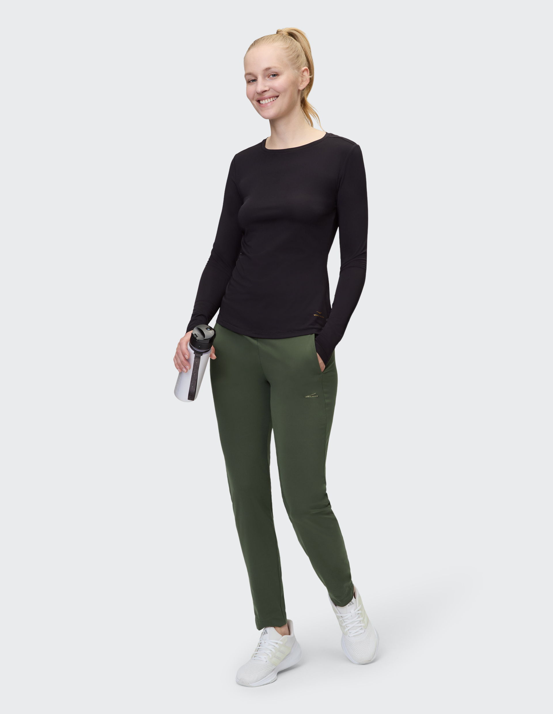 Eine Person trägt ein schwarzes langärmeliges Shirt, grüne Hose und weiße Turnschuhe, stehend vor grauem Hintergrund.