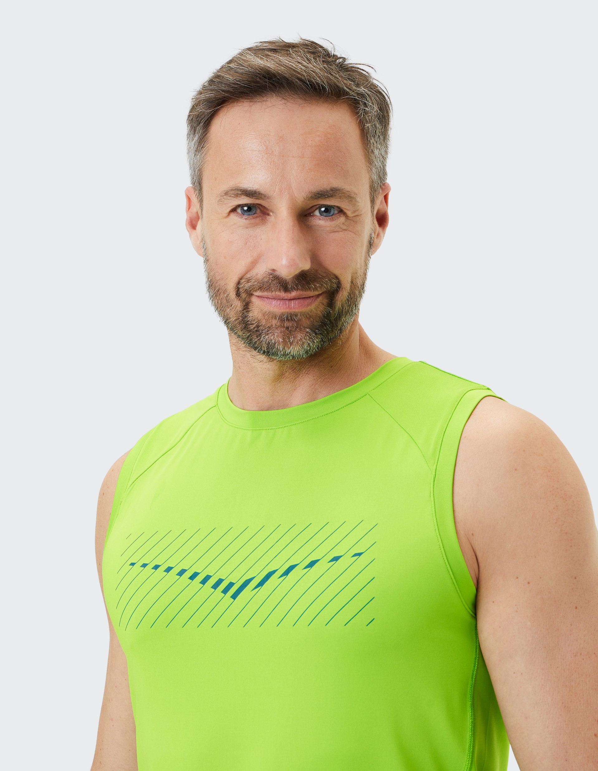 Mann mit grauen Haaren und Bart in neon-grünem ärmellosen Shirt mit dunkelblauem Muster lächelt freundlich vor einem hellen Hintergrund.
