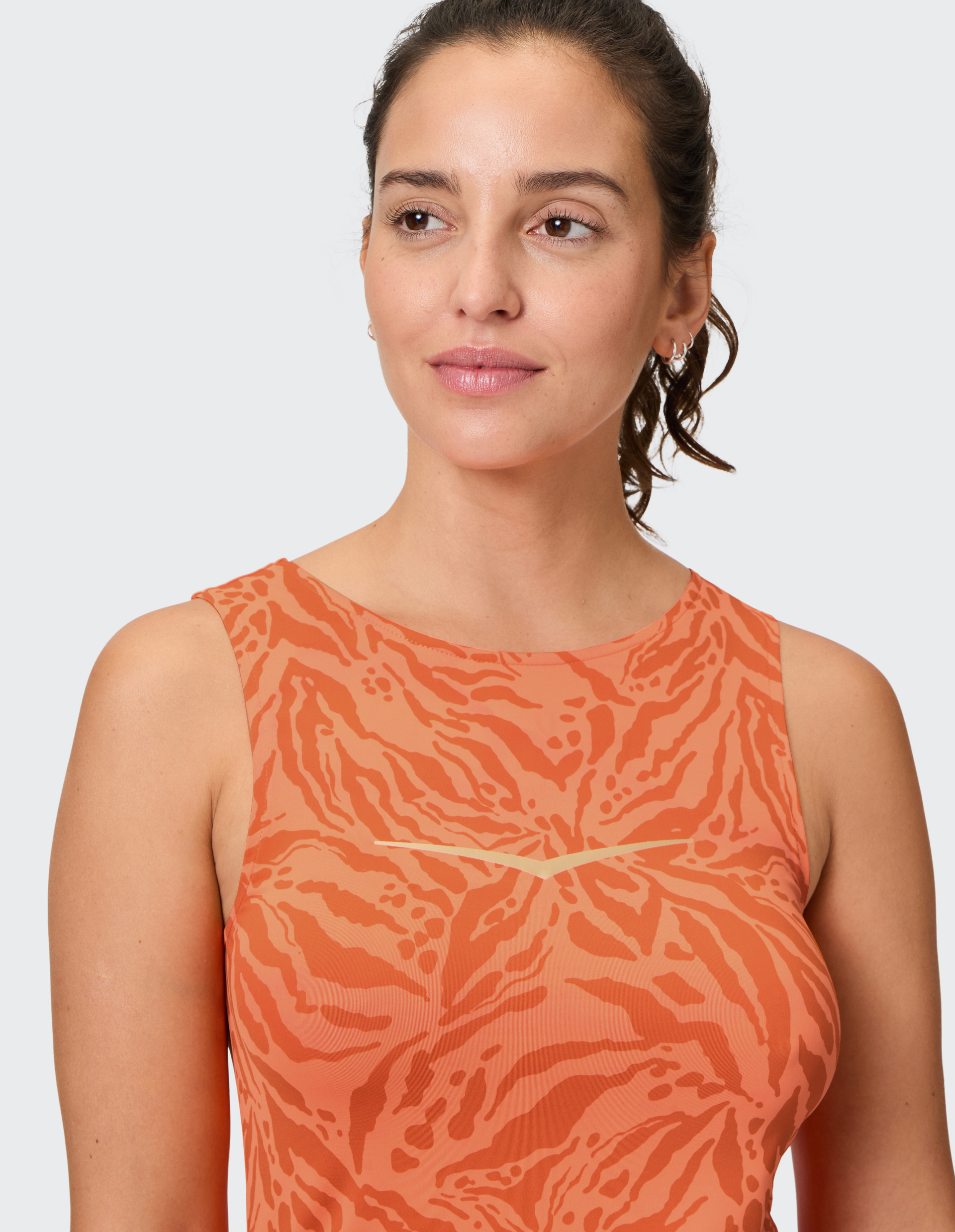 Frau mit lockigem Haar trägt ärmelloses, orange-gemustertes Oberteil vor hellem Hintergrund und schaut zur Seite.