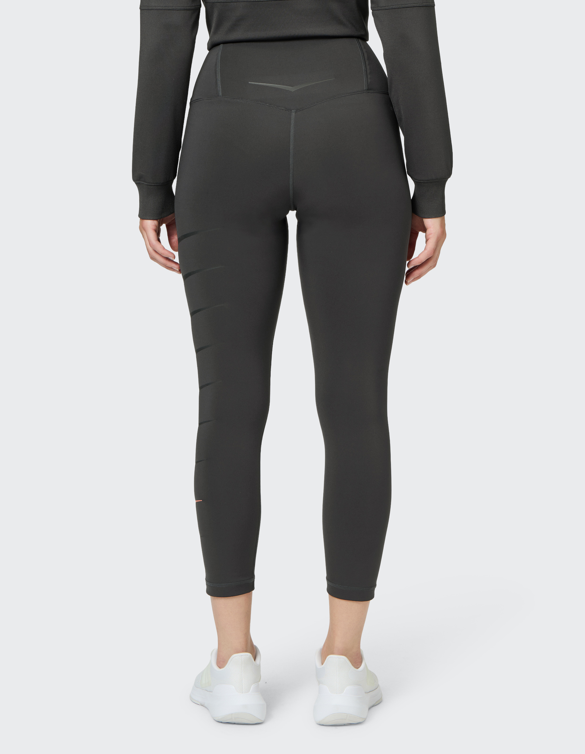 Rückansicht einer Person in dunklen Leggings und weißem Sneaker auf hellem Hintergrund, mit sichtbaren Nähten und strukturierten Details.