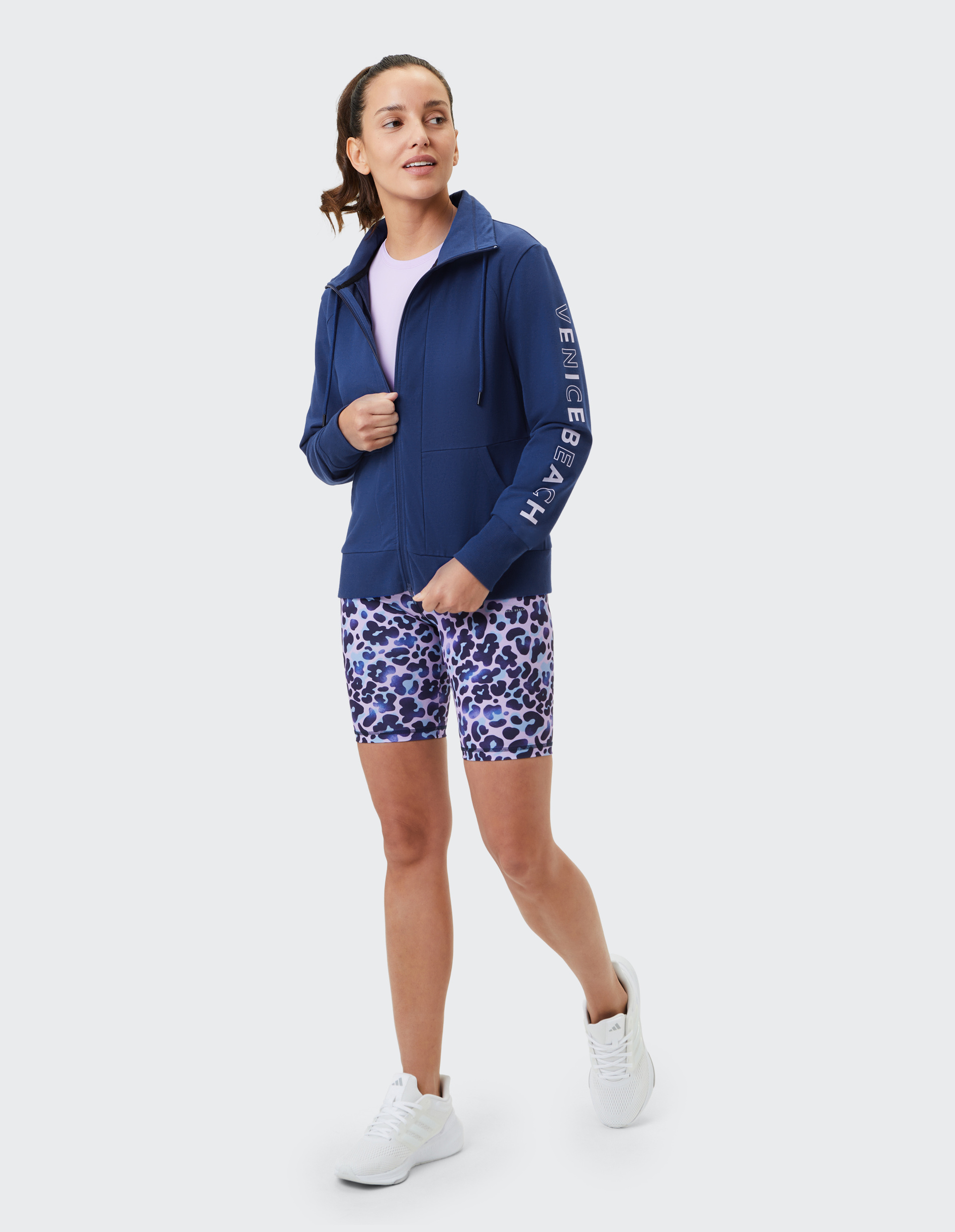 Frau in blauer Jacke mit "Venice Beach"-Schriftzug, lila Shirt und gemusterten Shorts geht lächelnd, weiße Sneaker ergänzen das Outfit.
