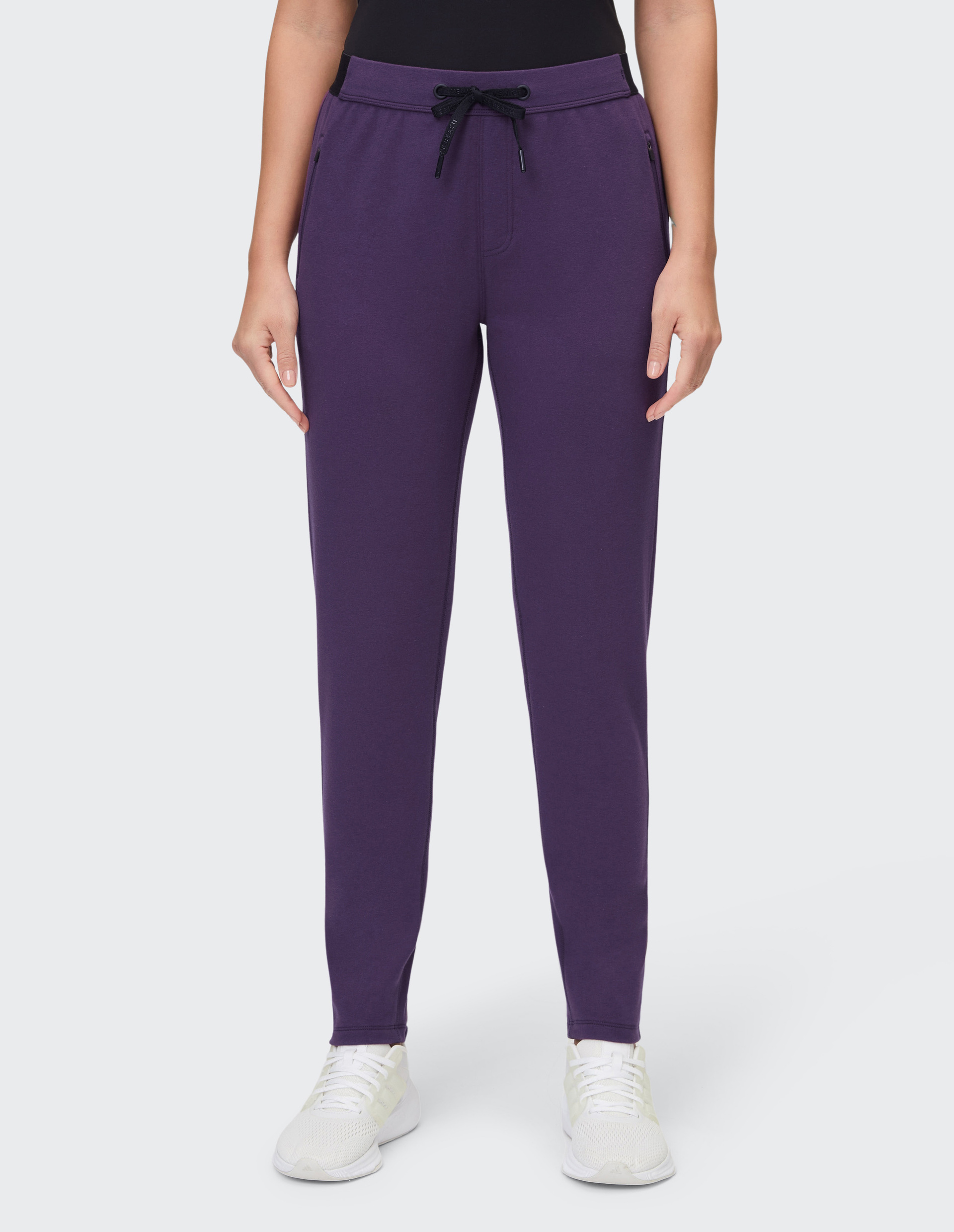 Frau trägt eine lilafarbene Jogginghose mit schwarzem Kordelzug und weißen Sneakern vor weißem Hintergrund.
