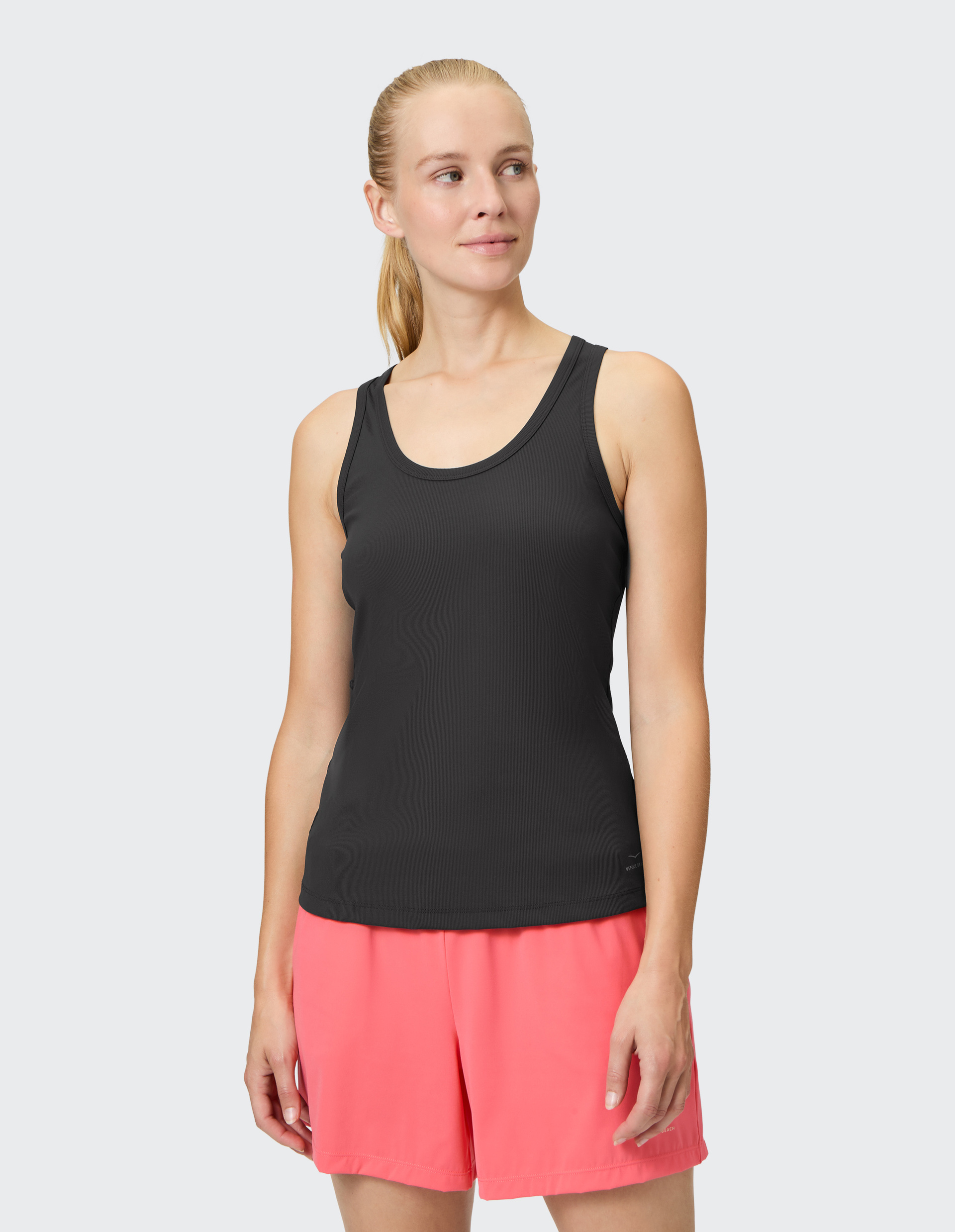 Frau in schwarzem Tanktop und korallroten Shorts schaut lächelnd zur Seite, vor einem hellgrauen Hintergrund stehend.