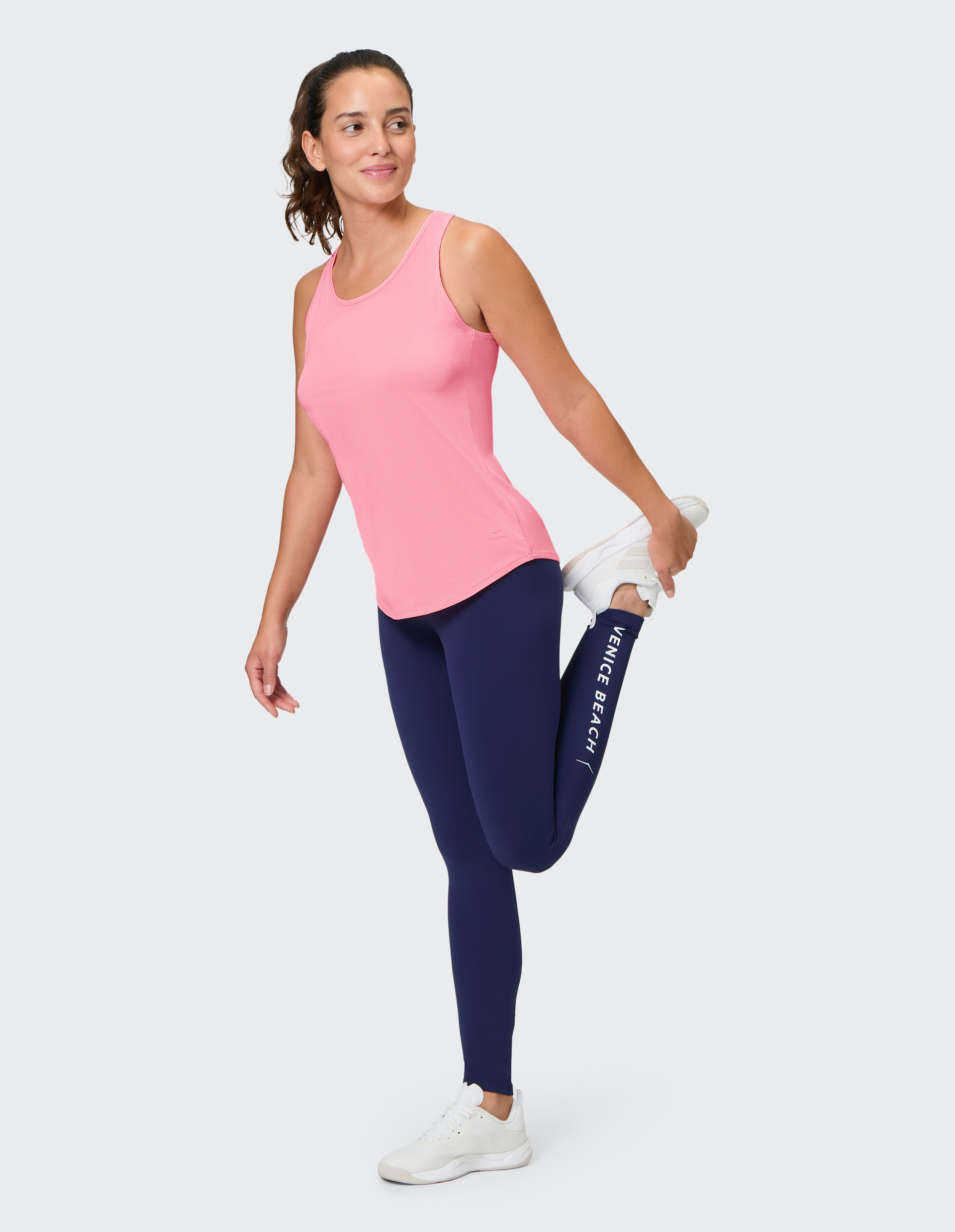 Frau in pinkem Tanktop und blauer Leggings dehnt ihr Bein, lächelt und trägt weiße Turnschuhe vor grauem Hintergrund.