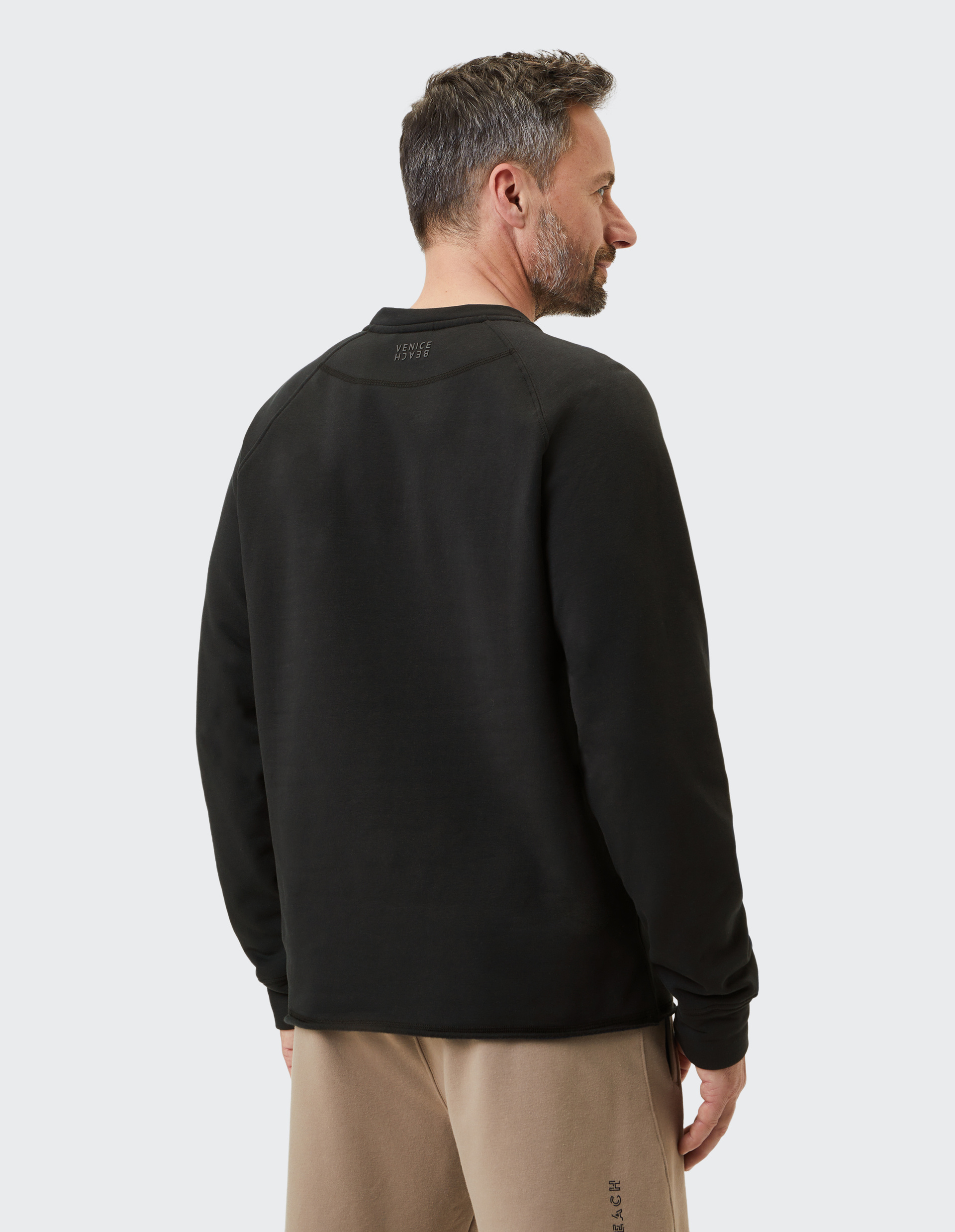 Ein Mann mit kurzem Haar und Bart trägt einen schwarzen Pullover und beige Hosen; er steht mit dem Rücken zur Kamera.
