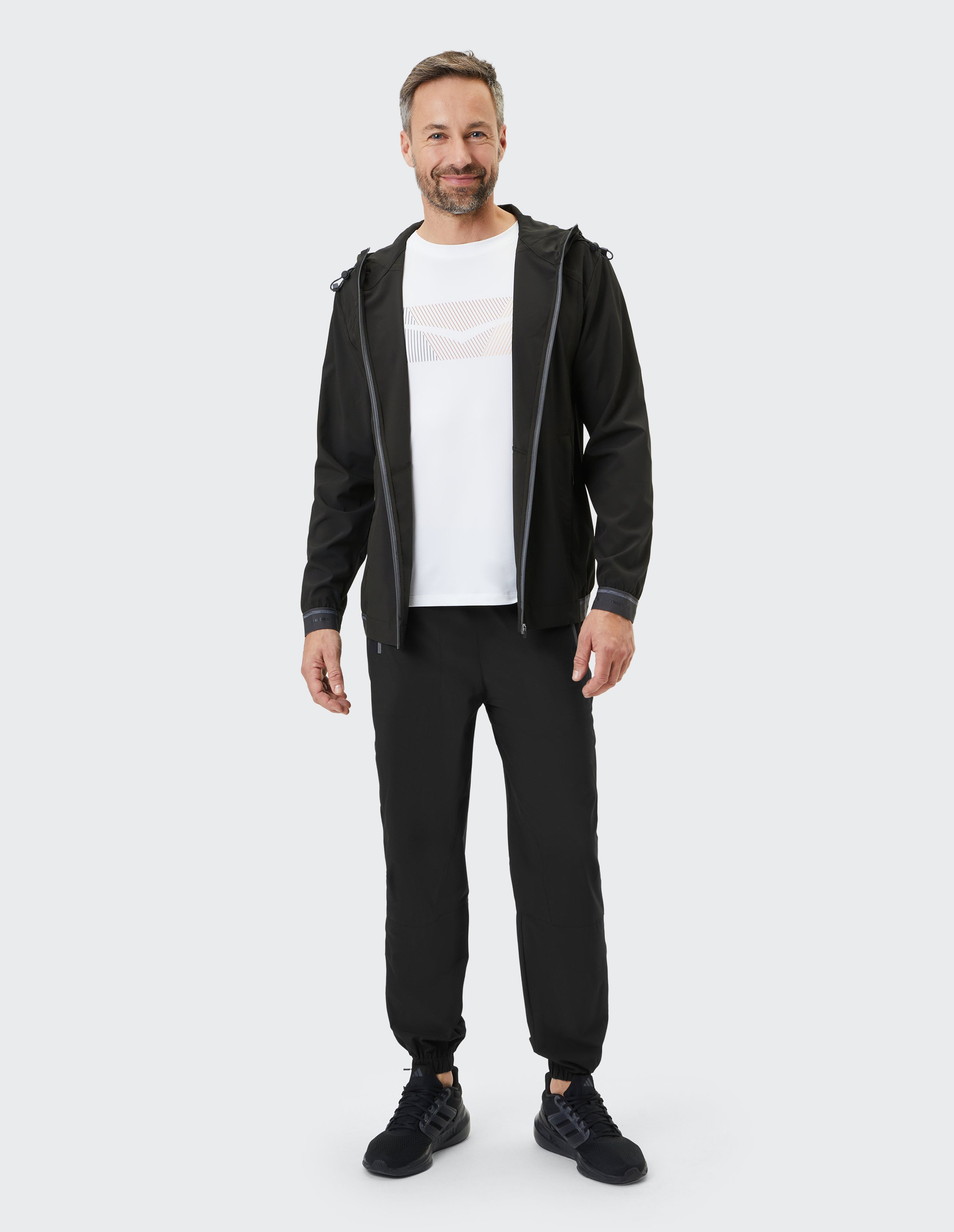 Mann lächelt in lässigem Outfit mit schwarzer Jacke und Hose, weißem Shirt und schwarzen Sneakers vor grauem Hintergrund.