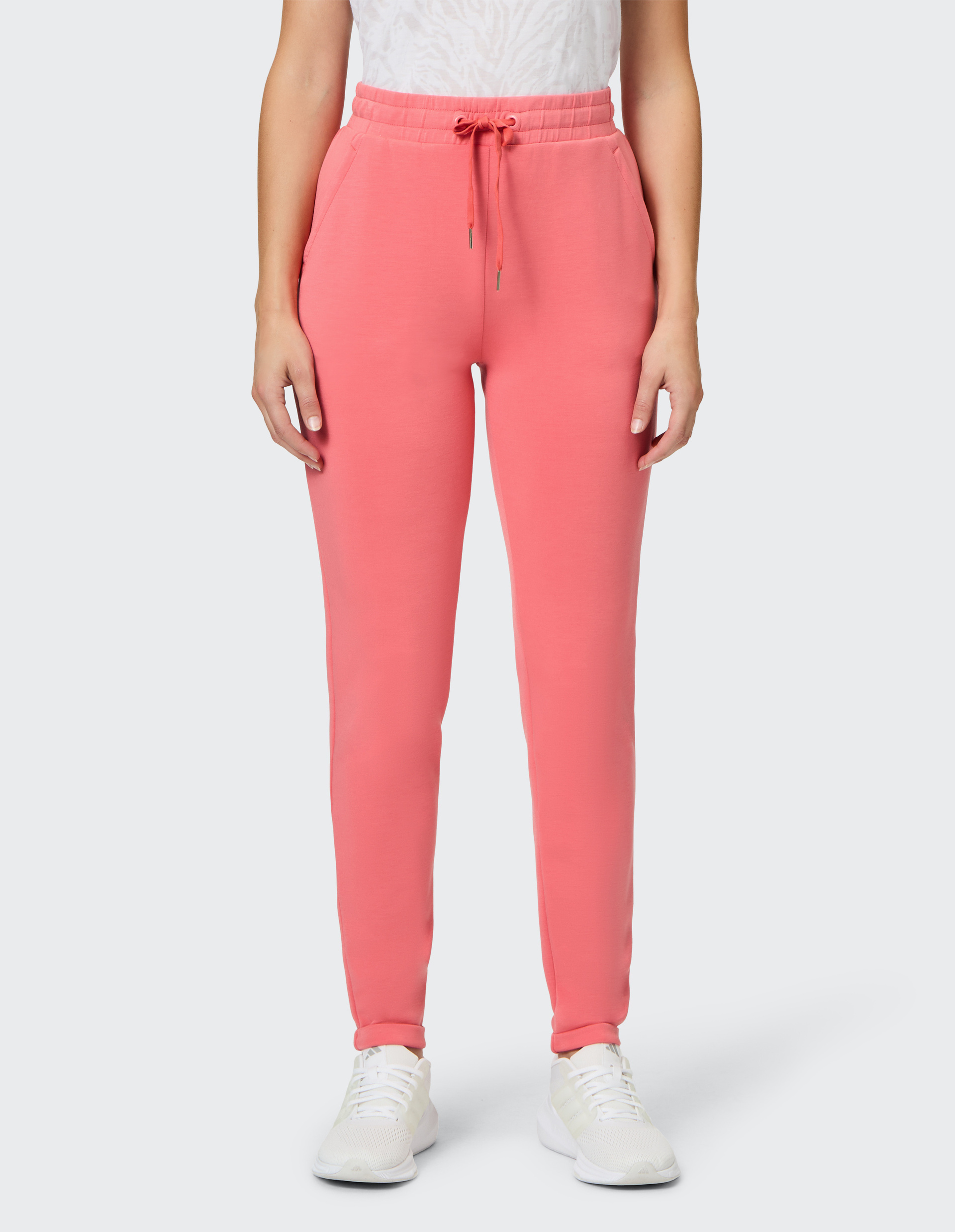 Person in korallfarbener Jogginghose mit Kordelzug und Seitentaschen, trägt weißen Sneaker auf hellem Hintergrund.