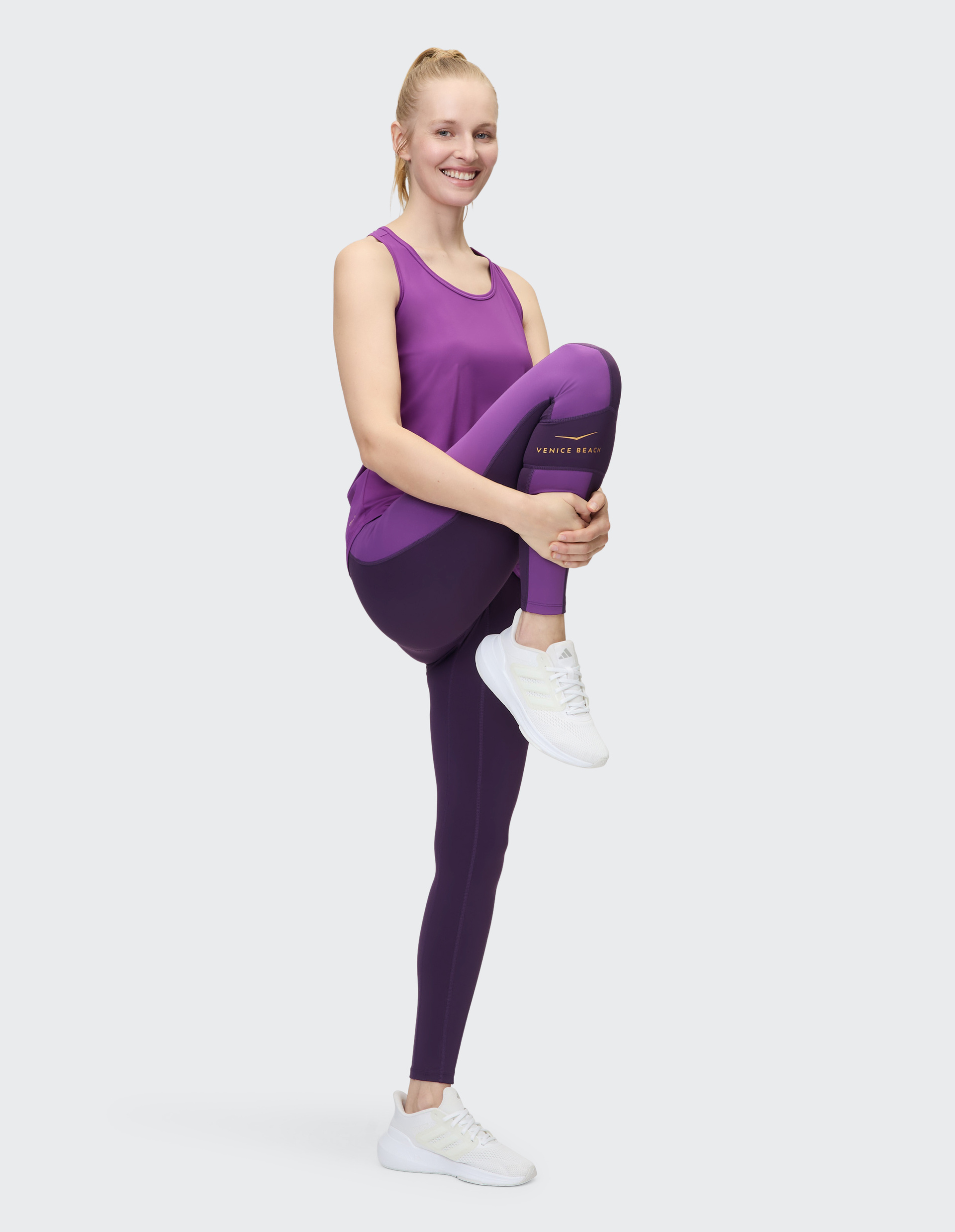 Eine lächelnde Frau in lila Sportkleidung macht eine Balanceübung und hält das Knie mit beiden Händen, auf weißem Hintergrund.