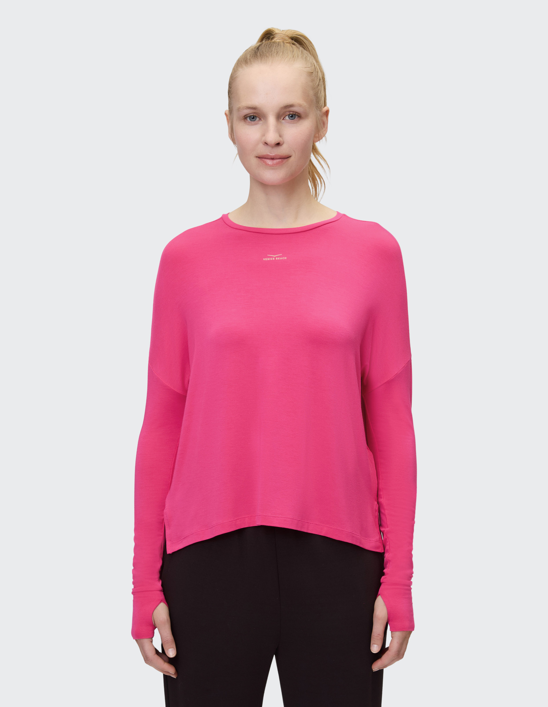 Frau trägt ein pinkes Langarmshirt und schwarze Hose, lächelt leicht und hat blonde Haare zu einem Pferdeschwanz gebunden.