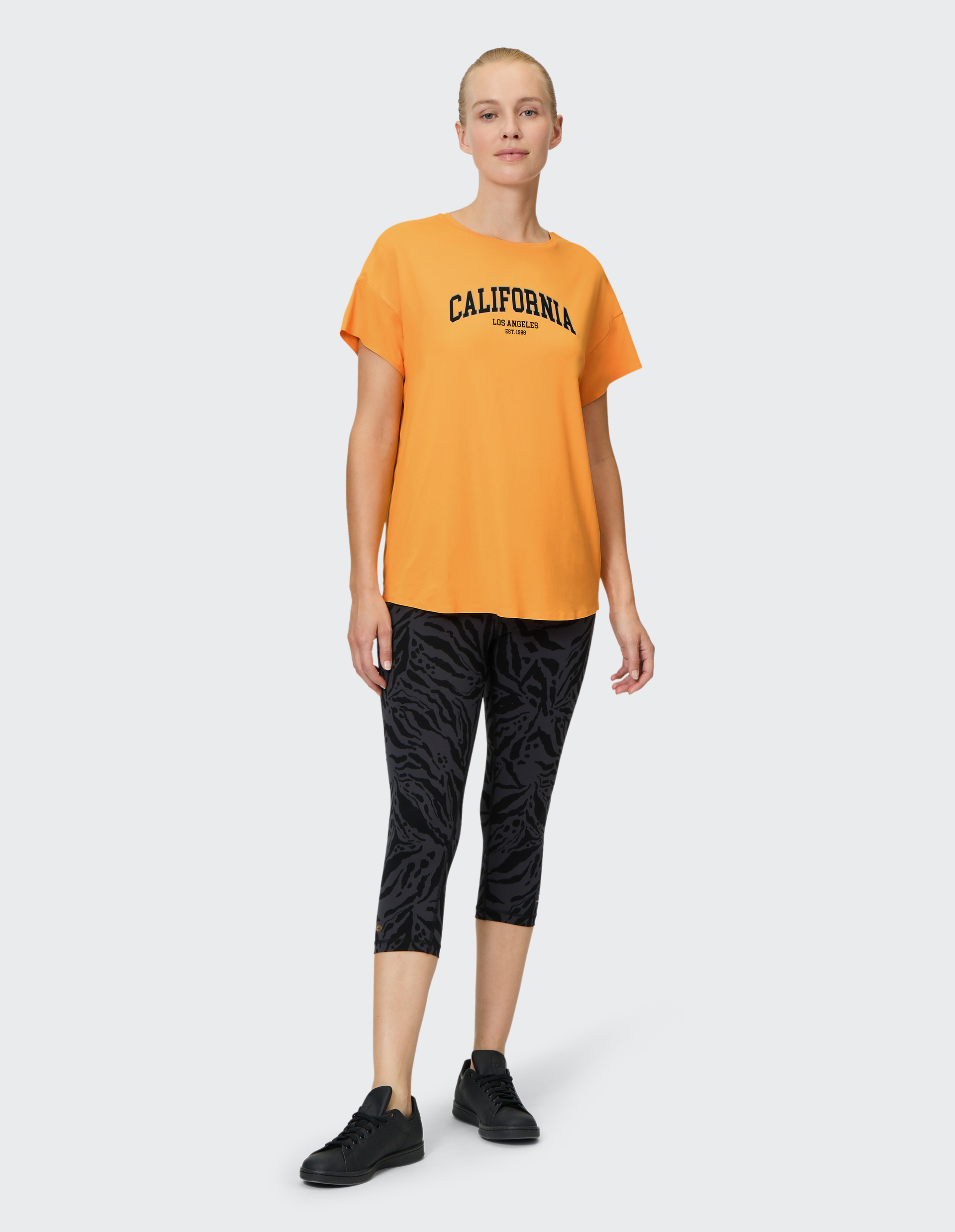 Frau in orangem "California"-Shirt und gemusterten Leggings, stehend, trägt schwarze Turnschuhe, lächelt leicht.