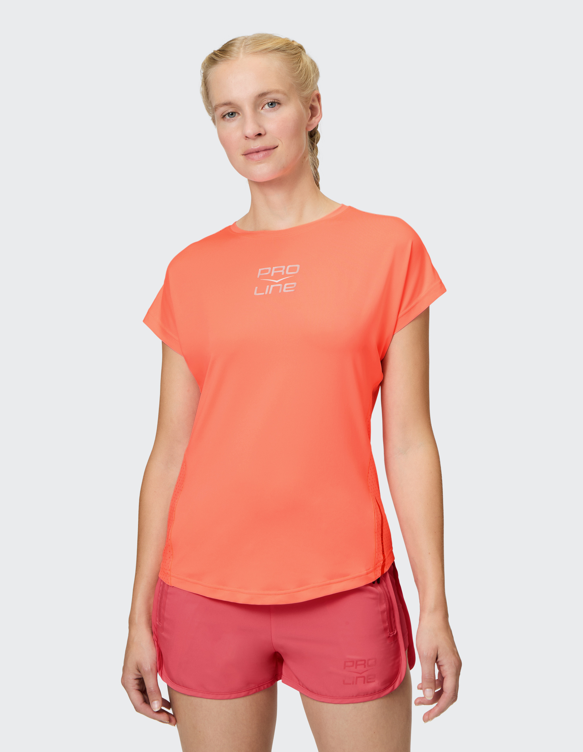 Frau mit blonden Zöpfen trägt ein leuchtend orangefarbenes T-Shirt und korallfarbene Shorts, beide mit "Pro Line" Logo, vor hellem Hintergrund.