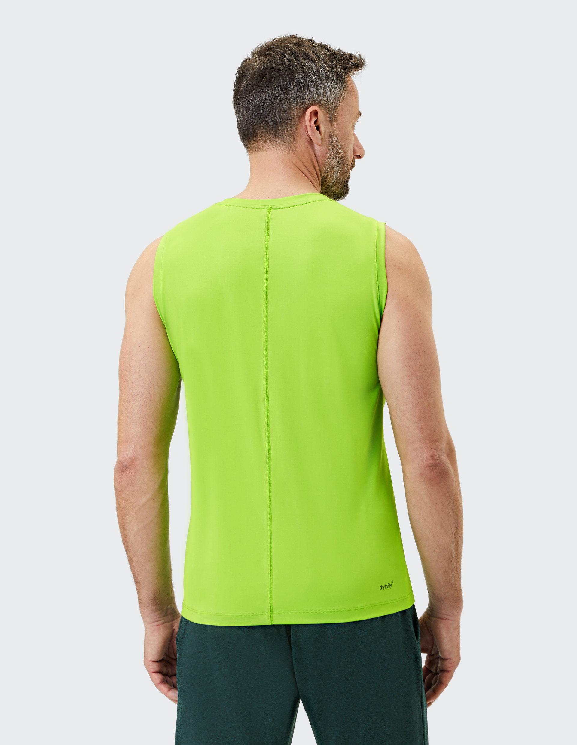 Ein Mann steht mit dem Rücken zur Kamera und trägt ein grünes ärmelloses Shirt und dunkelgrüne Hosen vor neutralem Hintergrund.