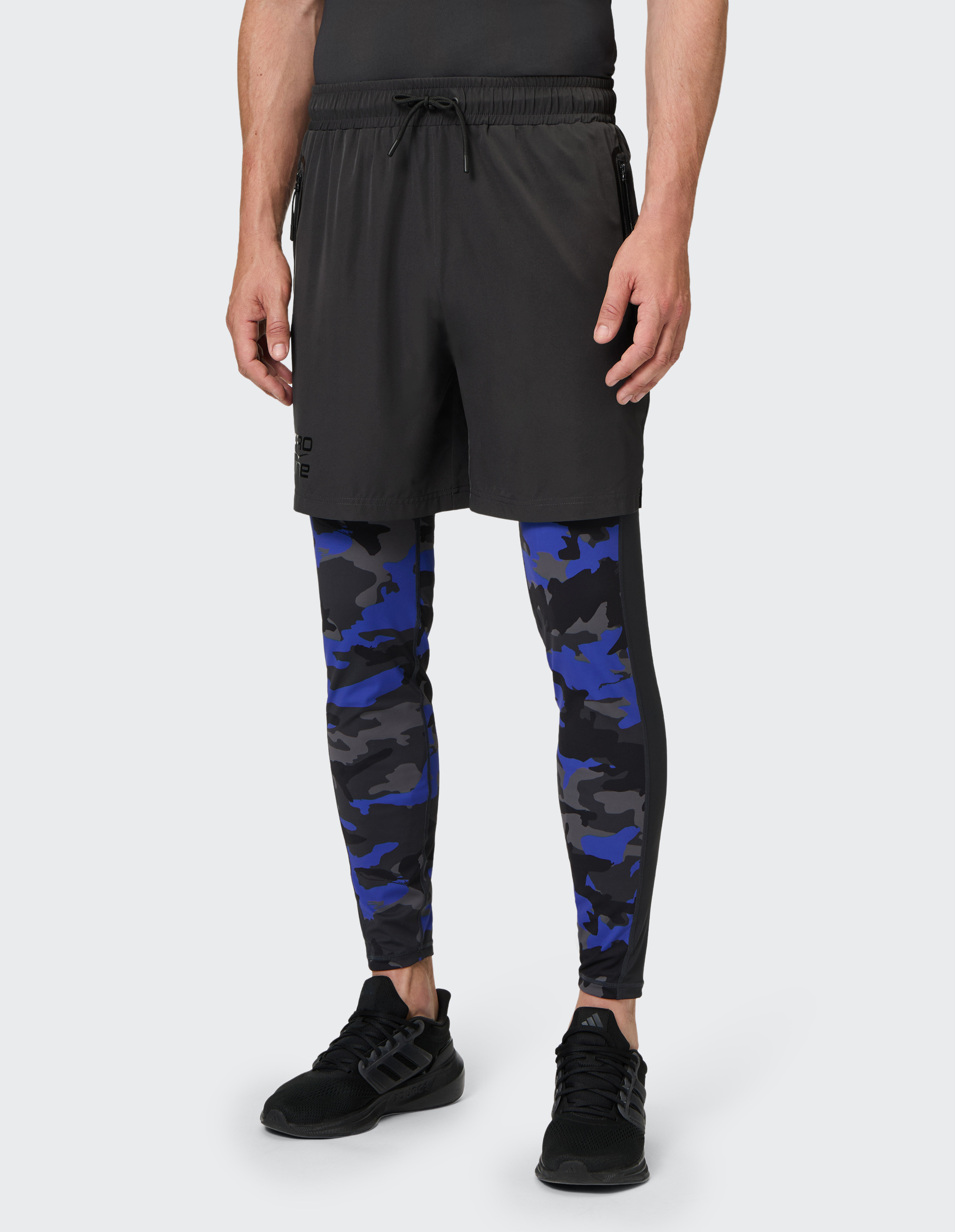 Mann in schwarzer Shorts mit blauer Camouflage-Leggings und schwarzen Sneakern auf grauem Hintergrund.