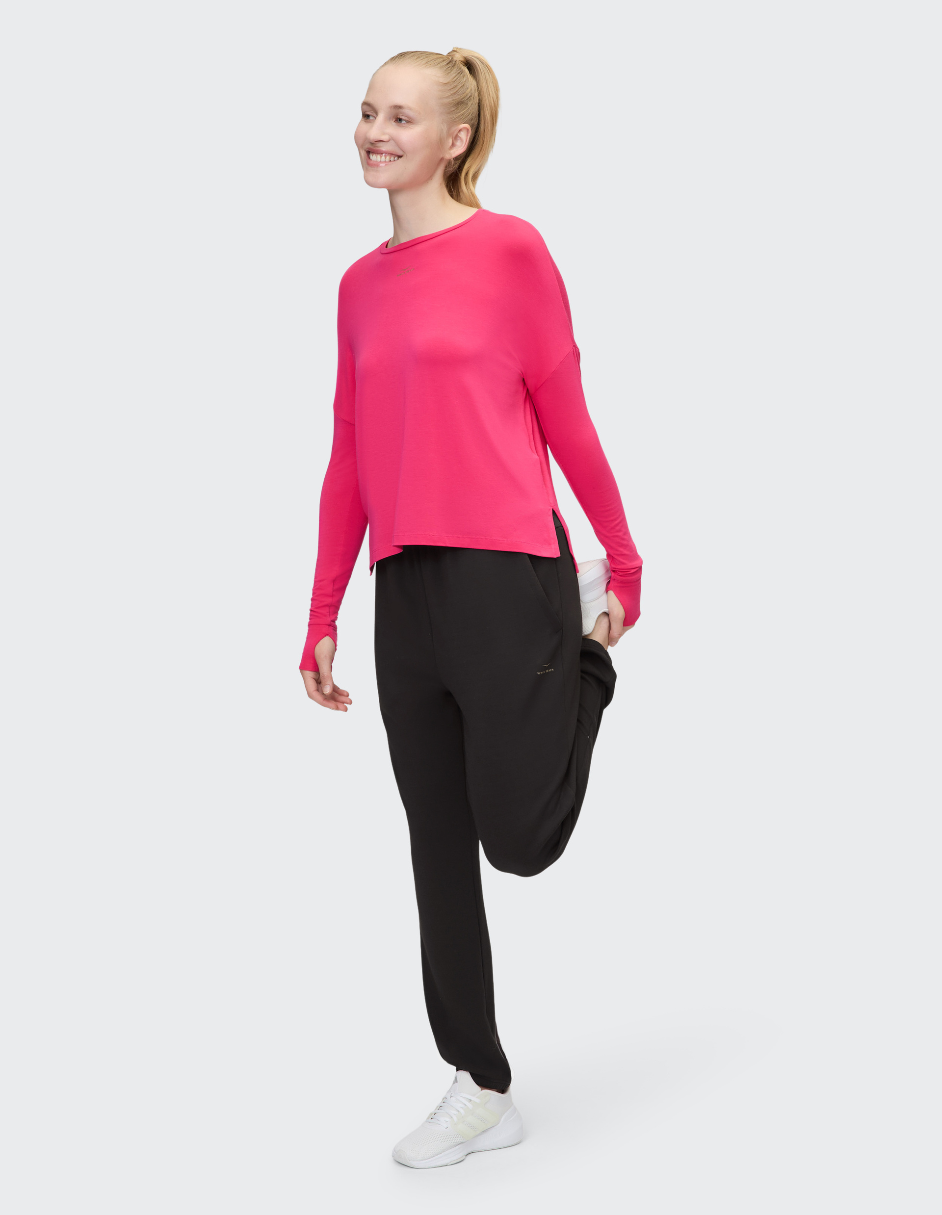 Eine Person trägt ein leuchtend pinkes langärmliges Oberteil, schwarze Jogginghose und weiße Turnschuhe vor einem weißen Hintergrund.