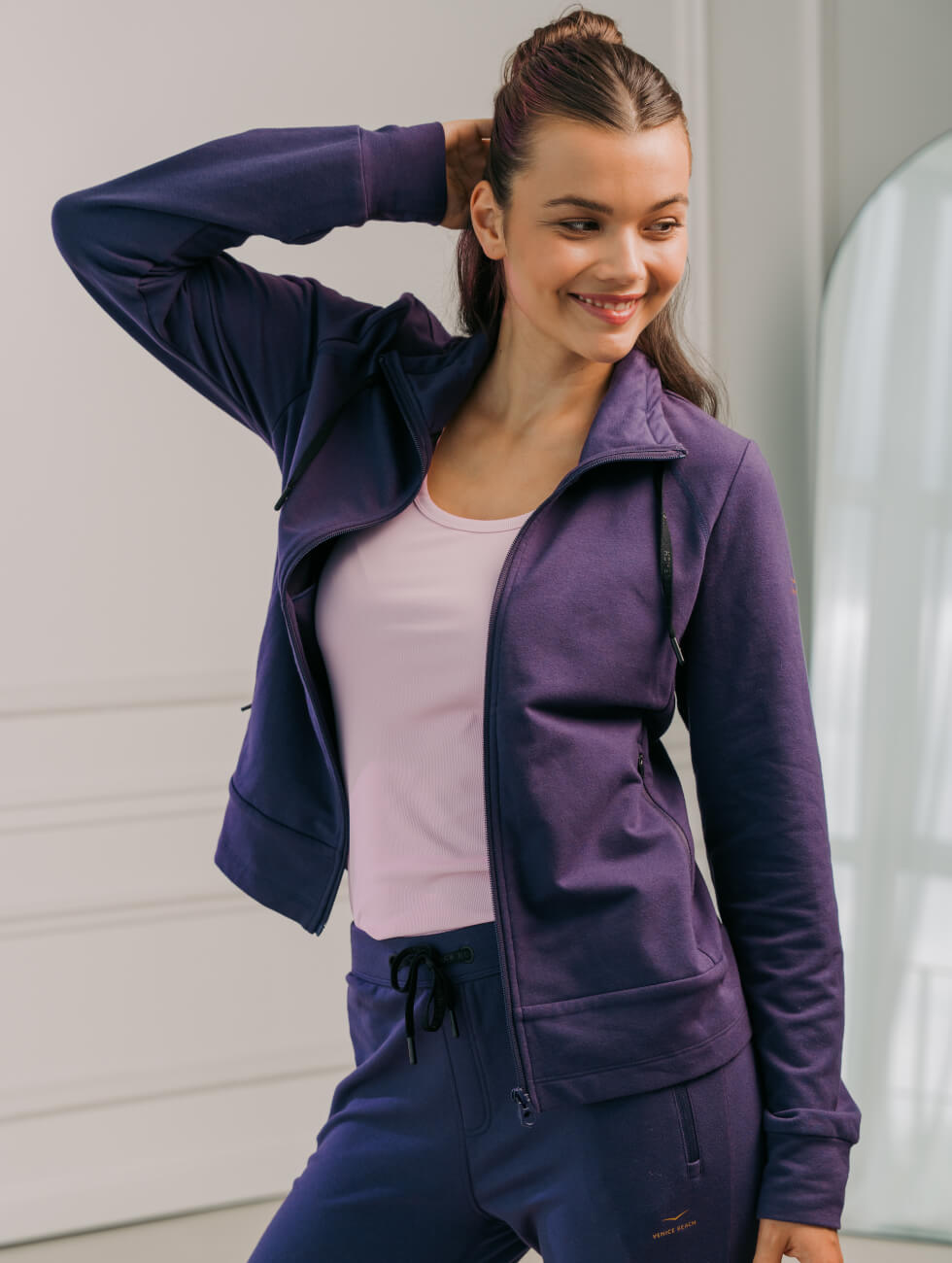 Frau in modischer Sportbekleidung mit lila Jogginganzug und blassrosa T-Shirt posiert im modernen, hellen Innenraum.