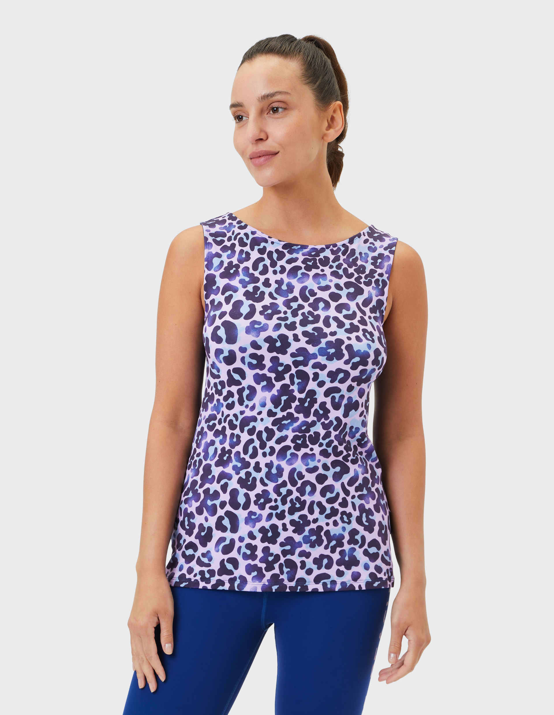 Frau trägt lila Top mit Leopardenmuster und blaue Leggings, lächelt leicht, Haar zu Pferdeschwanz gebunden, vor hellem Hintergrund.