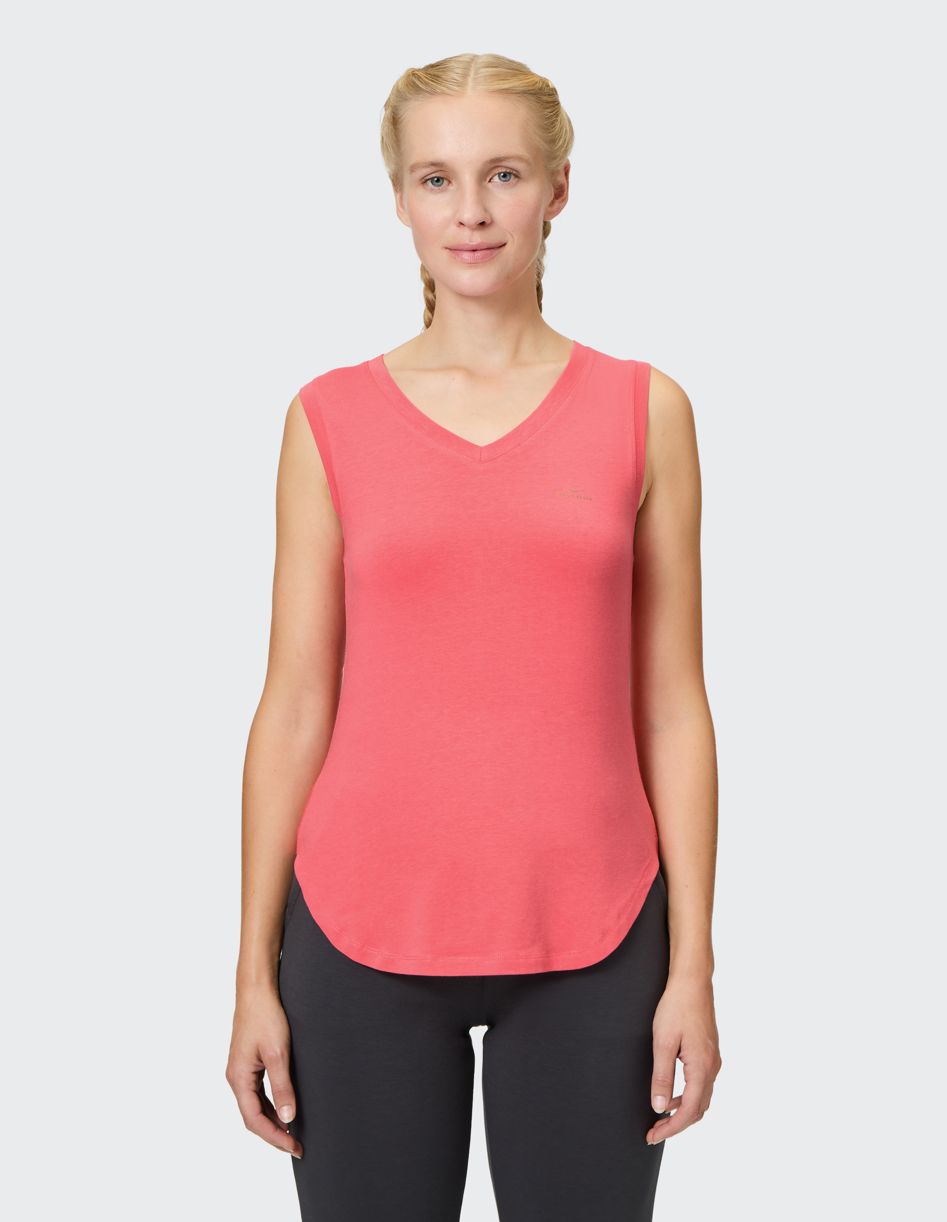 Eine Frau mit blonden Zöpfen trägt ein rotes ärmelloses Shirt und schwarze Leggings, vor einem hellgrauen Hintergrund stehend.