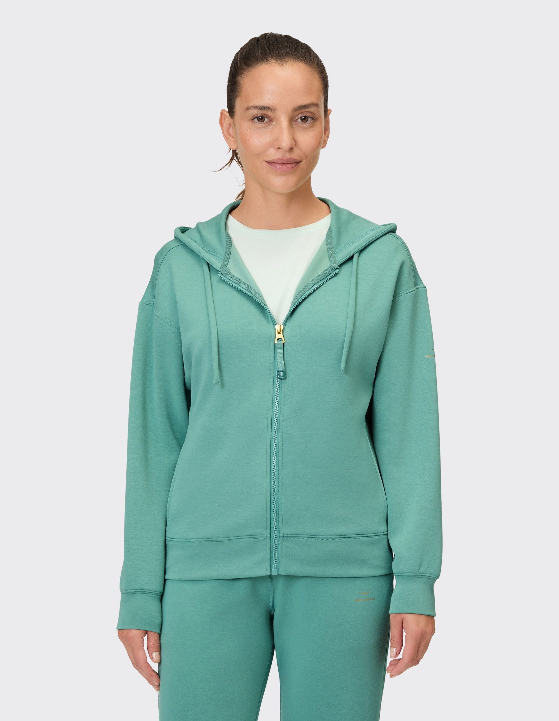 Eine Frau trägt einen petrolfarbenen Kapuzenpullover mit Reißverschluss und einer passenden Hose.