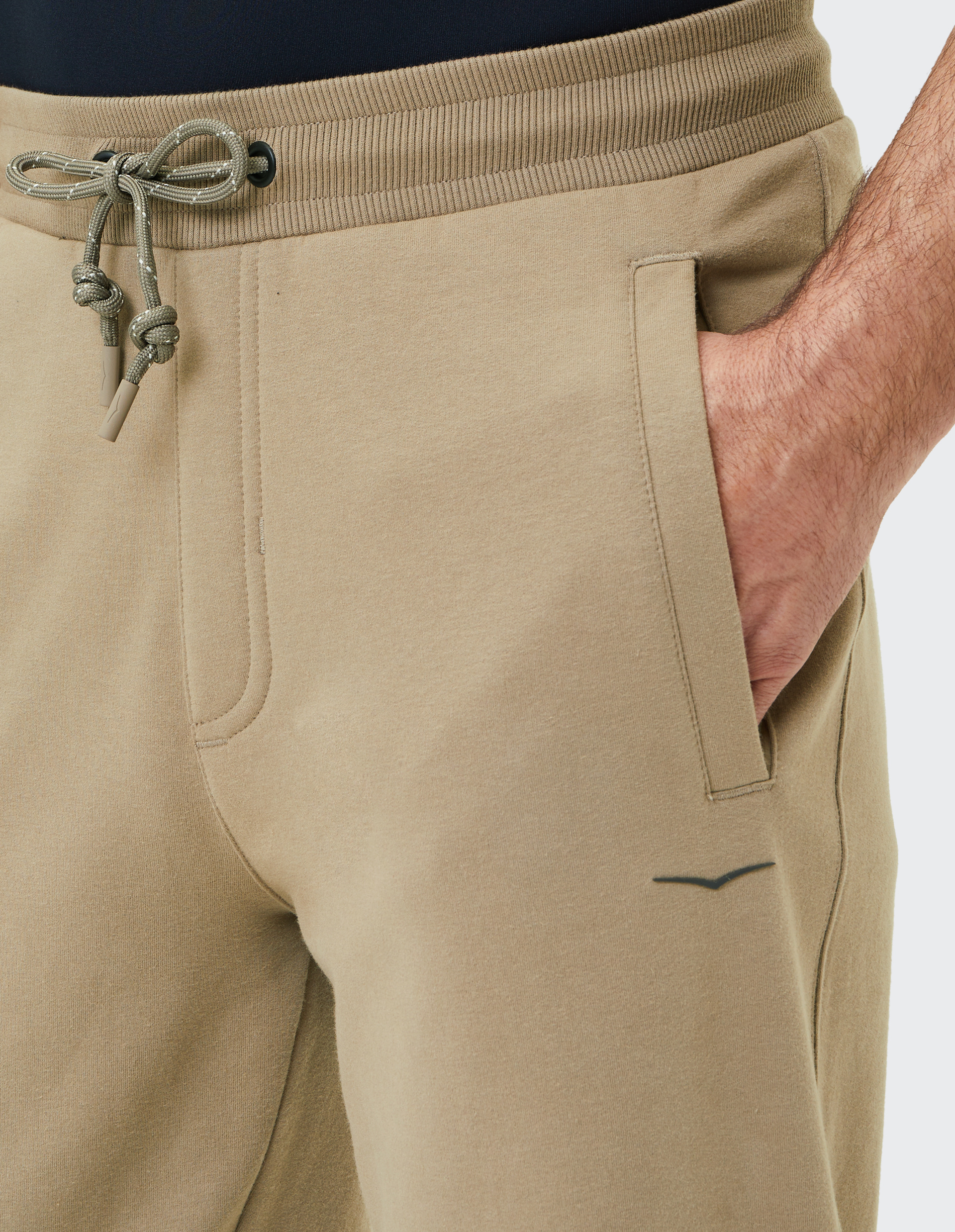 Beige Jogginghose mit elastischem Bund und Kordelzug, eine Hand steckt in der seitlichen Tasche; dezentes Logo vorhanden.