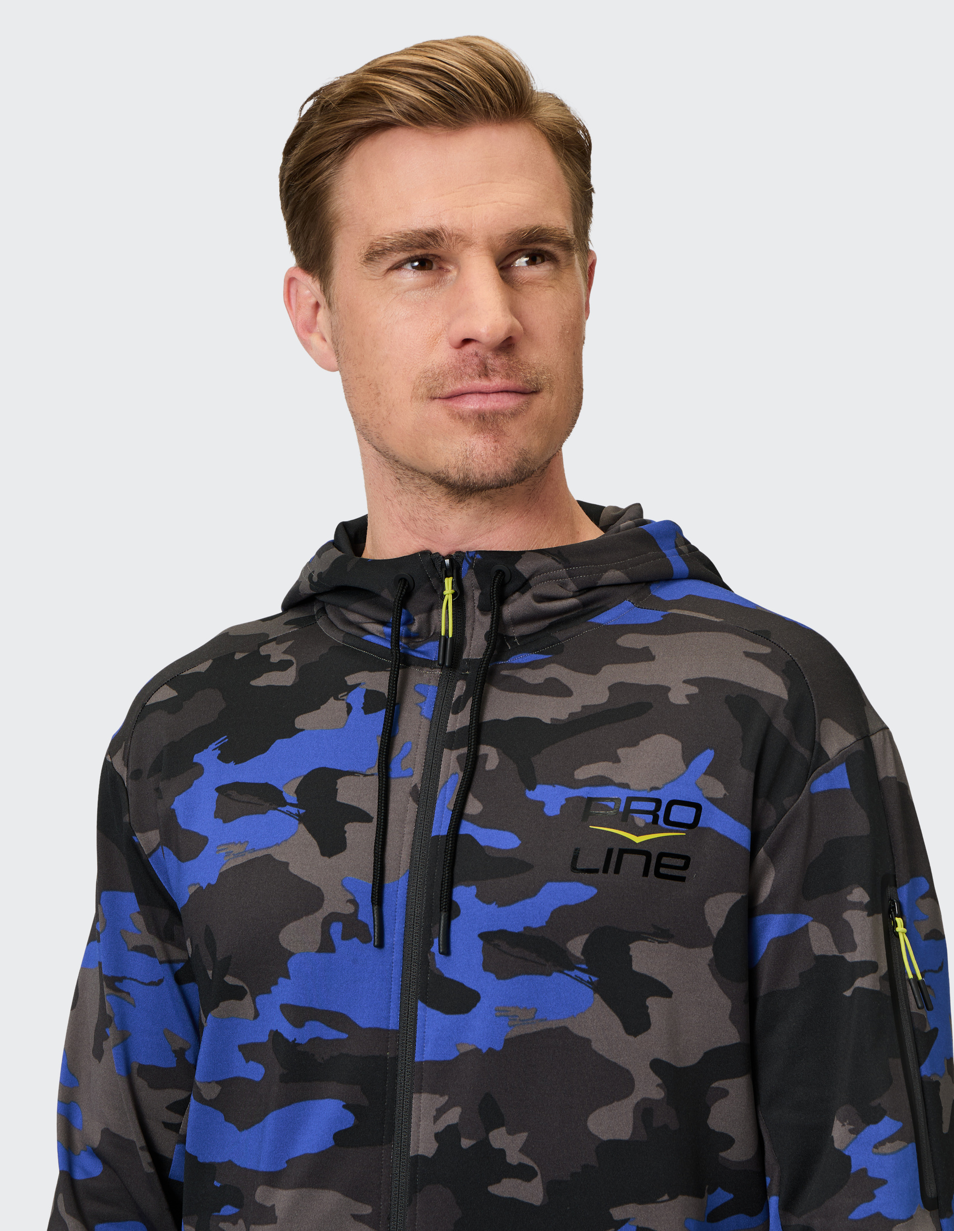Mann mit blonder Frisur trägt einen blauen und schwarzen Camouflage-Hoodie mit Pro Line-Aufschrift und Reißverschluss vor grauem Hintergrund.