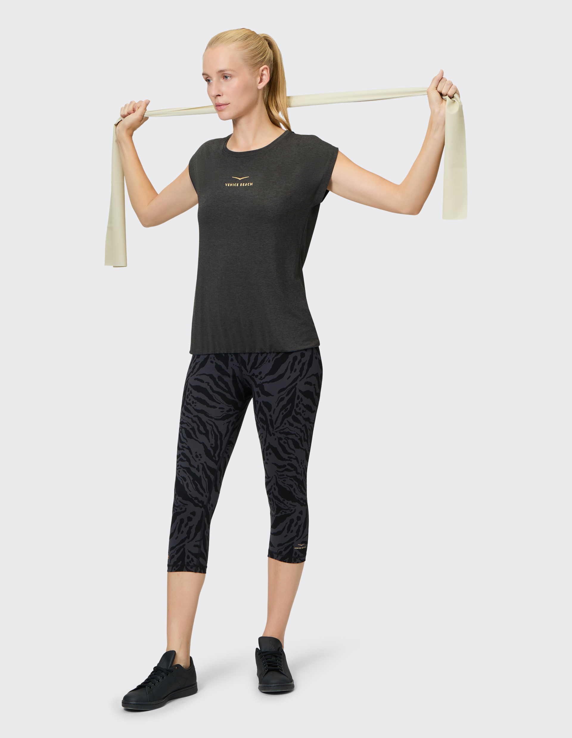 Frau in schwarzem Sportoutfit mit elastischem Band über Schultern; trägt schwarzes Shirt, gemusterte Leggings und schwarze Sneaker.