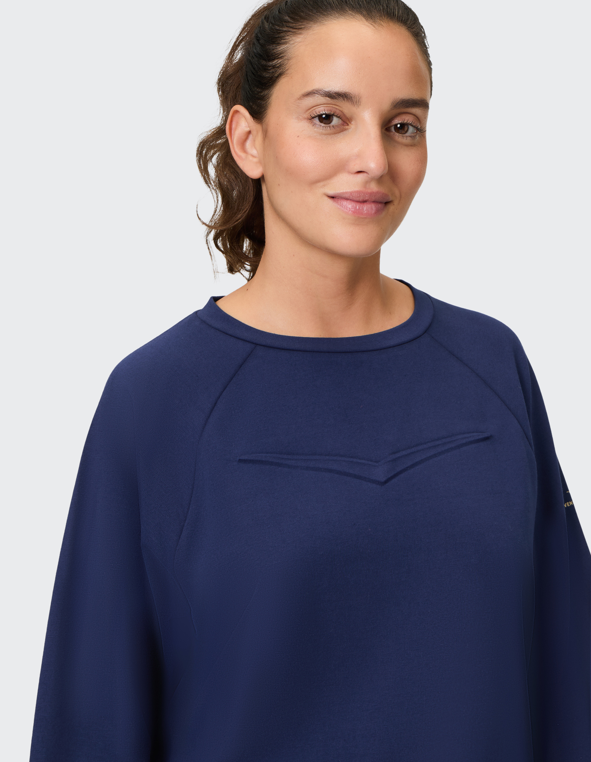Eine Frau mit zurückgebundenem Haar trägt einen dunkelblauen Pullover und lächelt leicht vor einem hellgrauen Hintergrund.