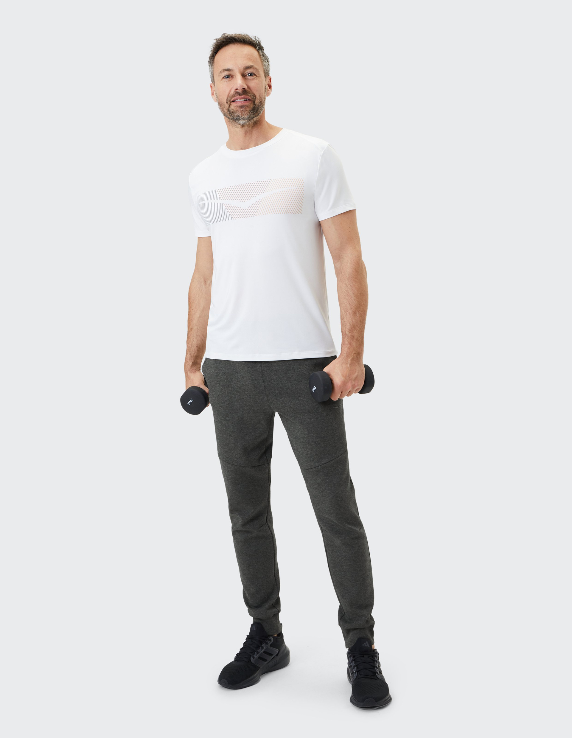 Ein Mann in einem weißen T-Shirt und grauer Jogginghose hält Hanteln, steht aufrecht und lächelt in die Kamera.