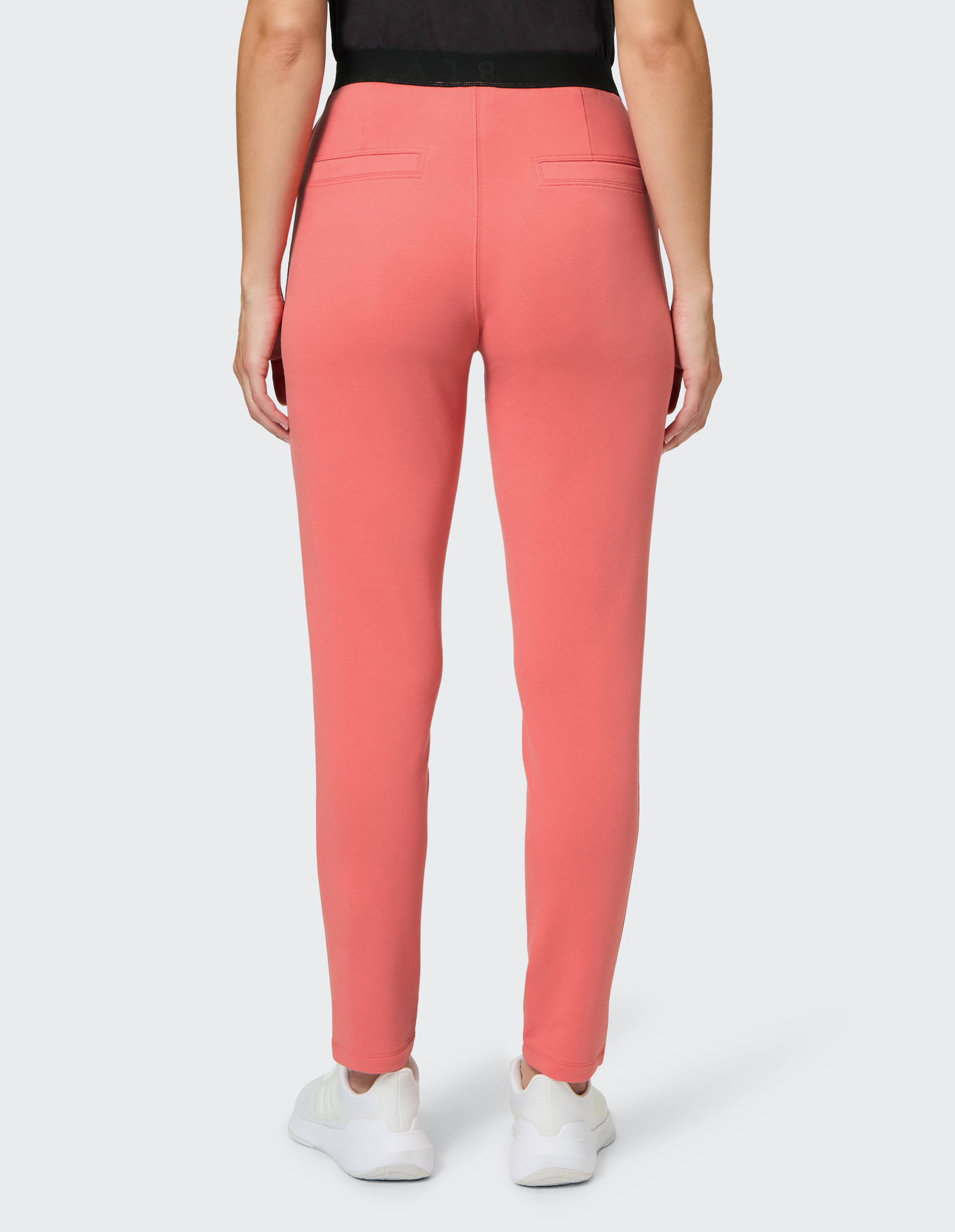 Frau trägt korallrote Hose mit zwei Gesäßtaschen und weiße Sneakers, steht mit dem Rücken zur Kamera vor grauem Hintergrund.