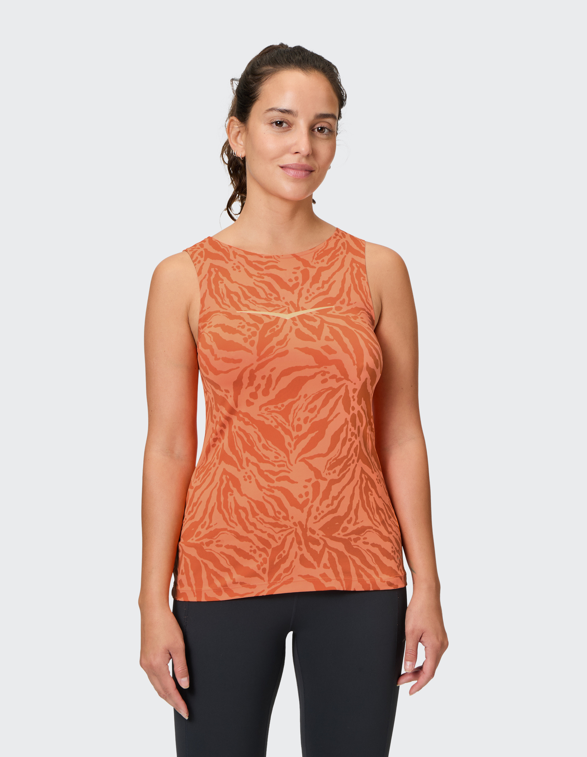 Eine Frau trägt ein orangefarbenes Tanktop mit abstraktem Muster und schwarze Hosen, vor einem schlichten weißen Hintergrund.