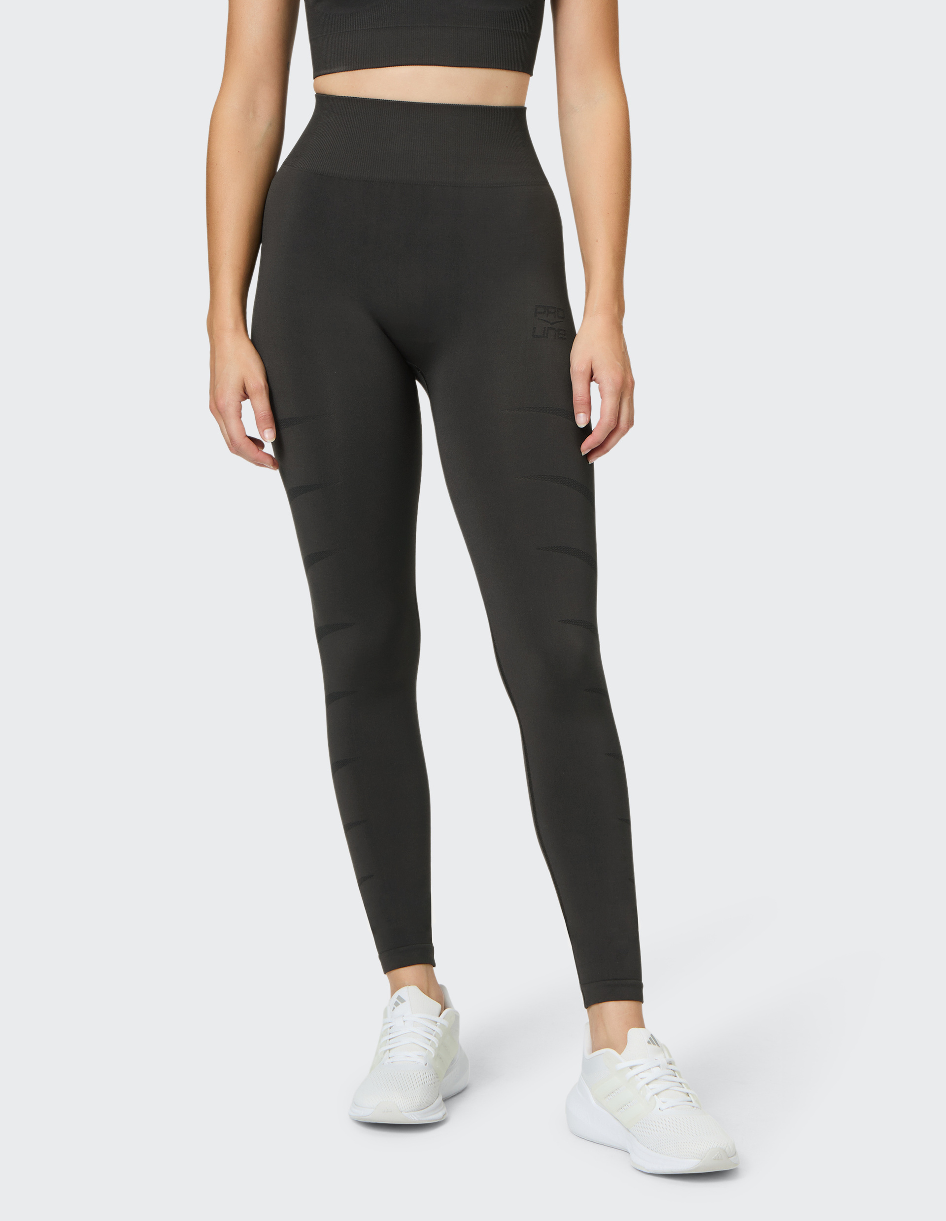 Frau trägt ein schwarzes Sport-Outfit mit Leggings und Top, kombiniert mit weißen Sportschuhen, vor einem hellgrauen Hintergrund.
