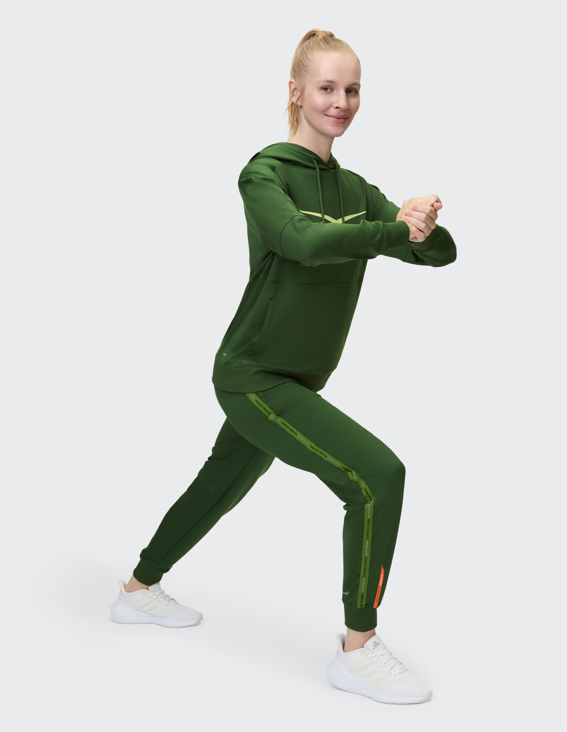 Frau in grünem Sportanzug macht Ausfallschritt, lächelt, trägt weiße Turnschuhe, auf hellem Hintergrund.