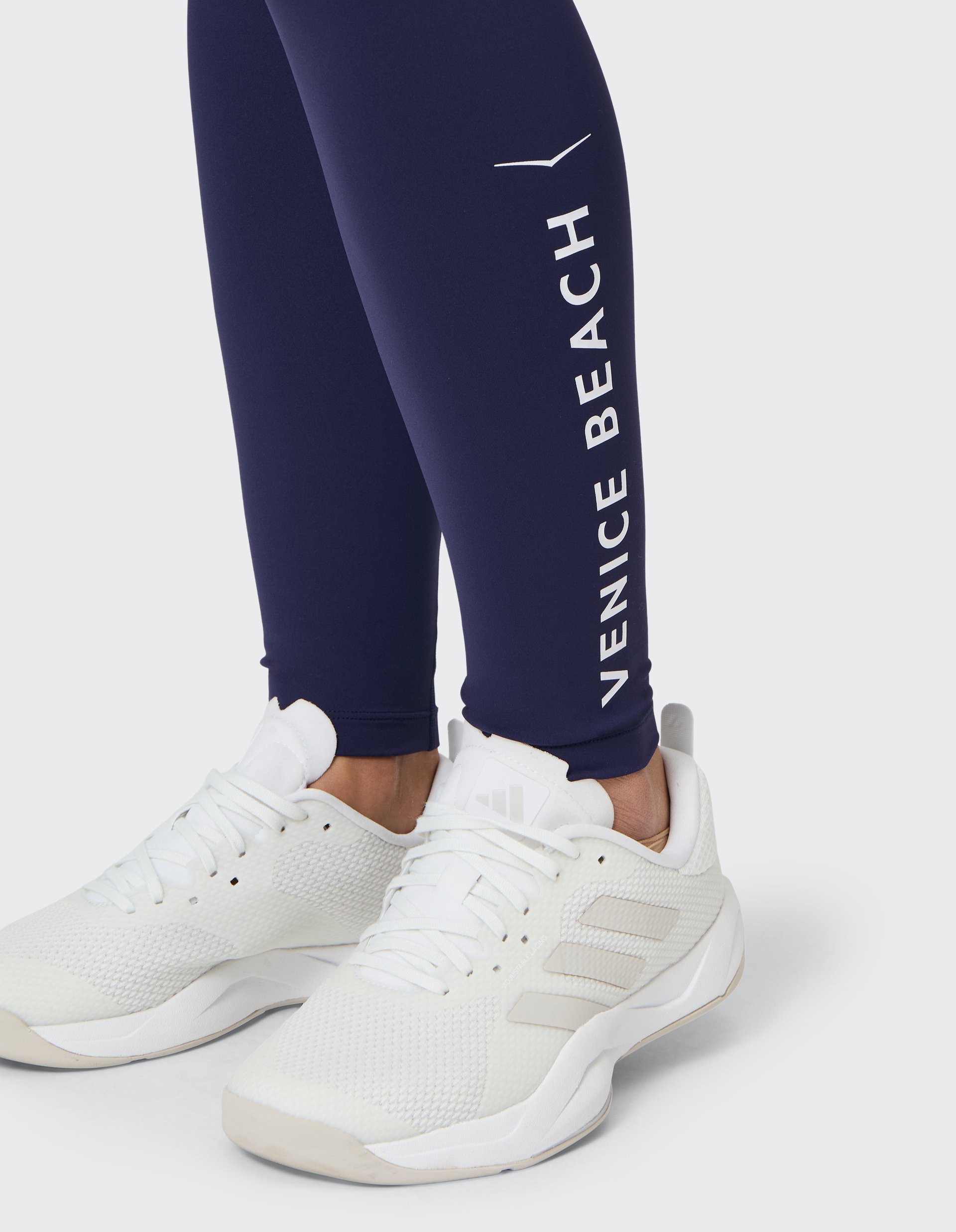 Weißer Sportschuh auf grauem Boden und dunkelblaue Leggings mit weißem "Venice Beach" Schriftzug an einem Bein.