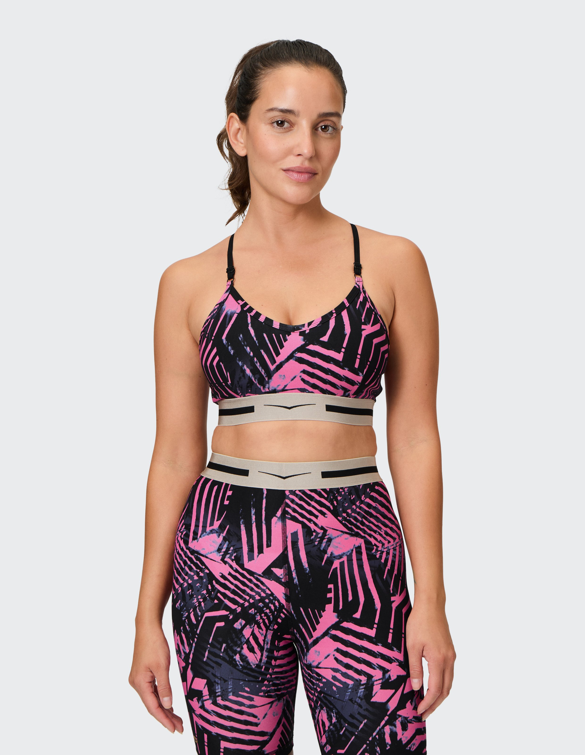 Frau in einem pink-schwarz gemusterten Sportoutfit mit Spaghettiträgern, stehend vor neutralem Hintergrund.
