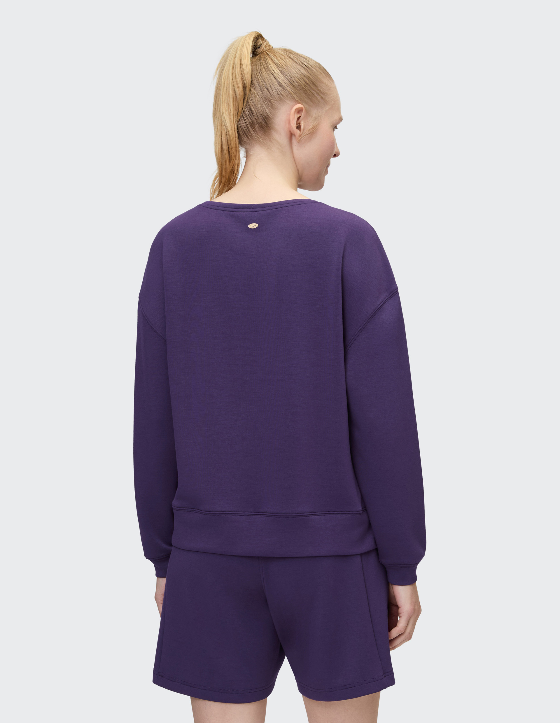 Frau mit blondem Pferdeschwanz von hinten in lila Sweatshirt und Shorts vor grauem Hintergrund.