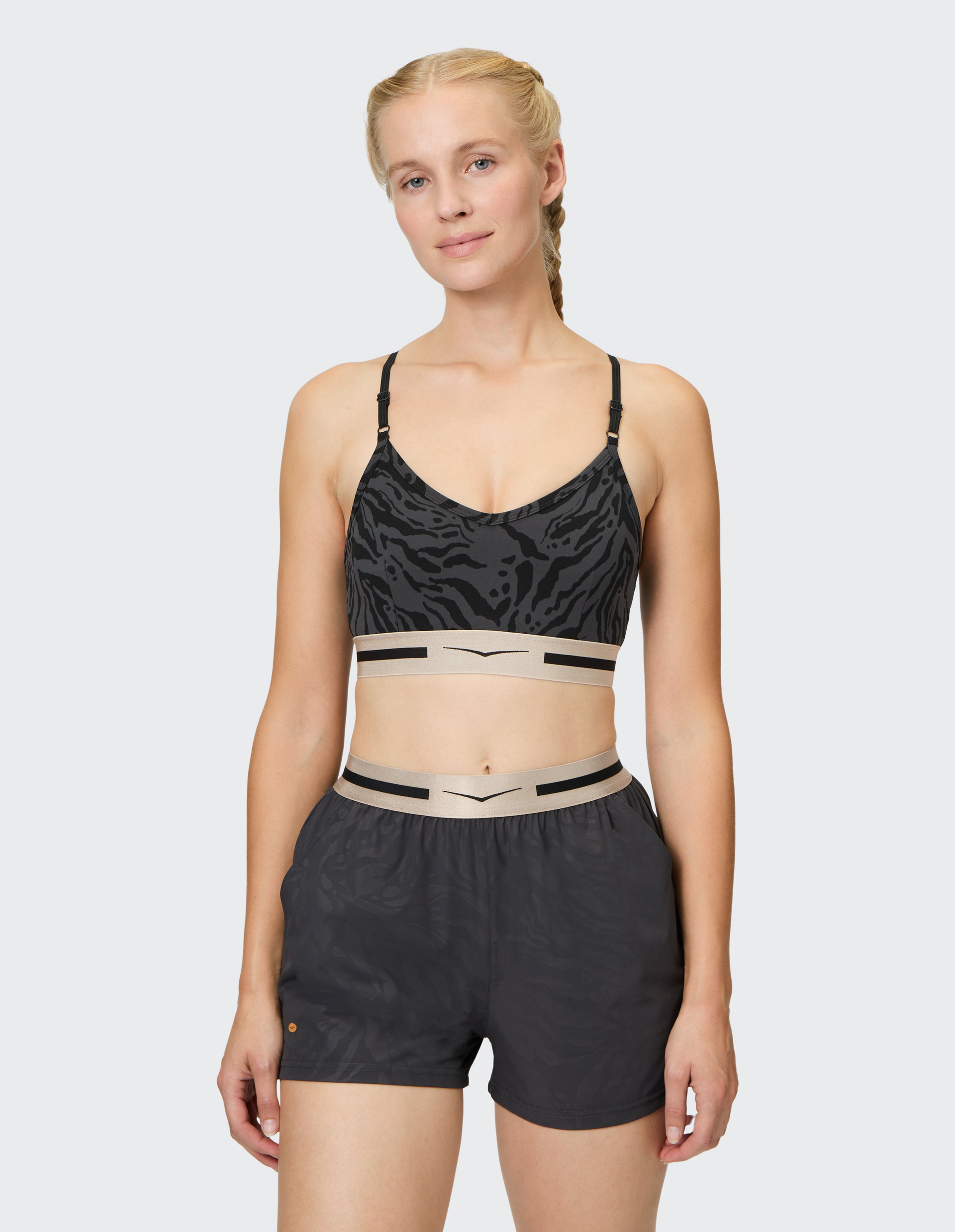 Frau trägt einen schwarz gemusterten Sport-BH und Shorts, lächelt leicht, mit geflochtenem Haar, vor neutralem Hintergrund.