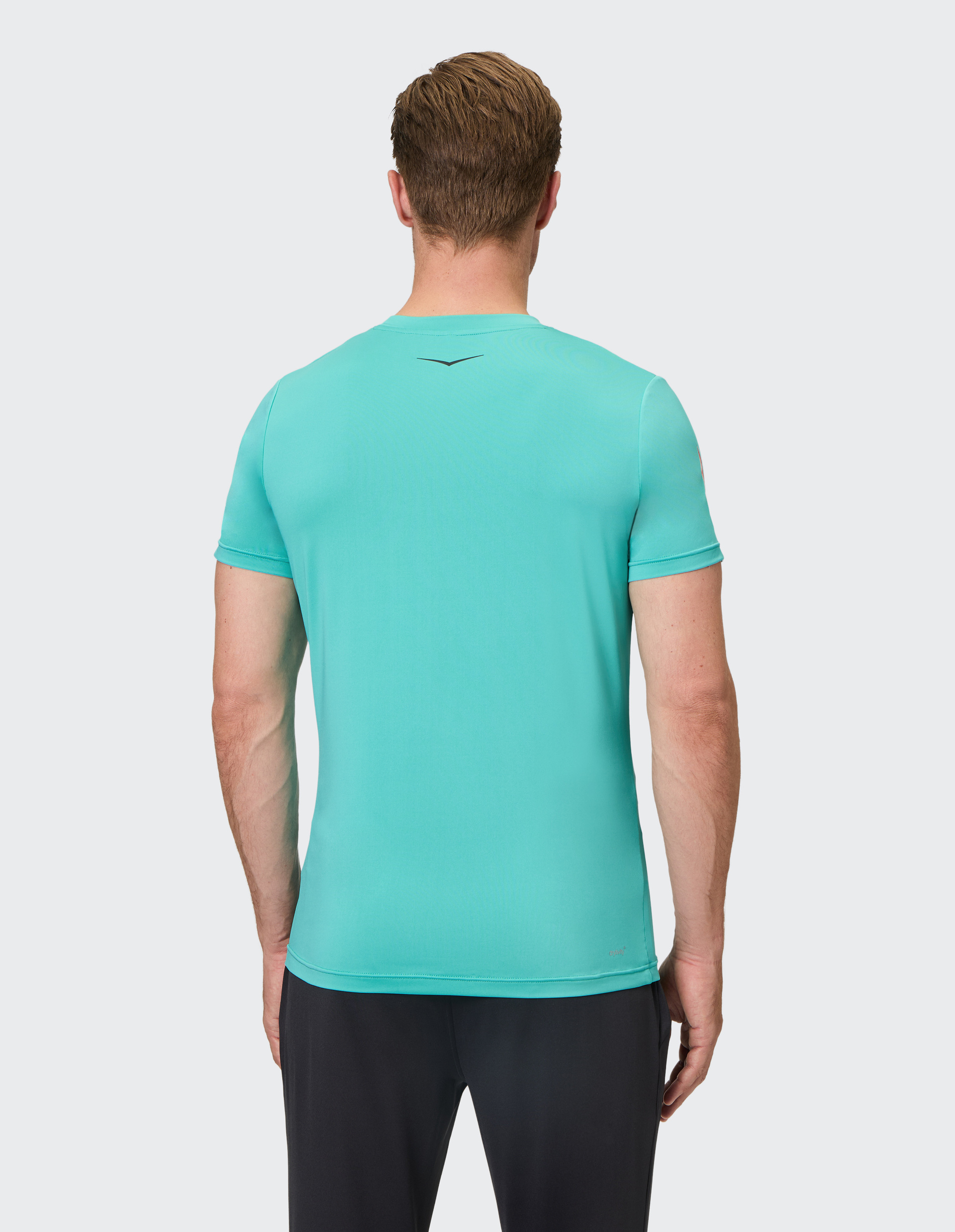 Ein Mann von hinten in einem türkisfarbenen T-Shirt und schwarzen Hosen, vor einem hellgrauen Hintergrund.