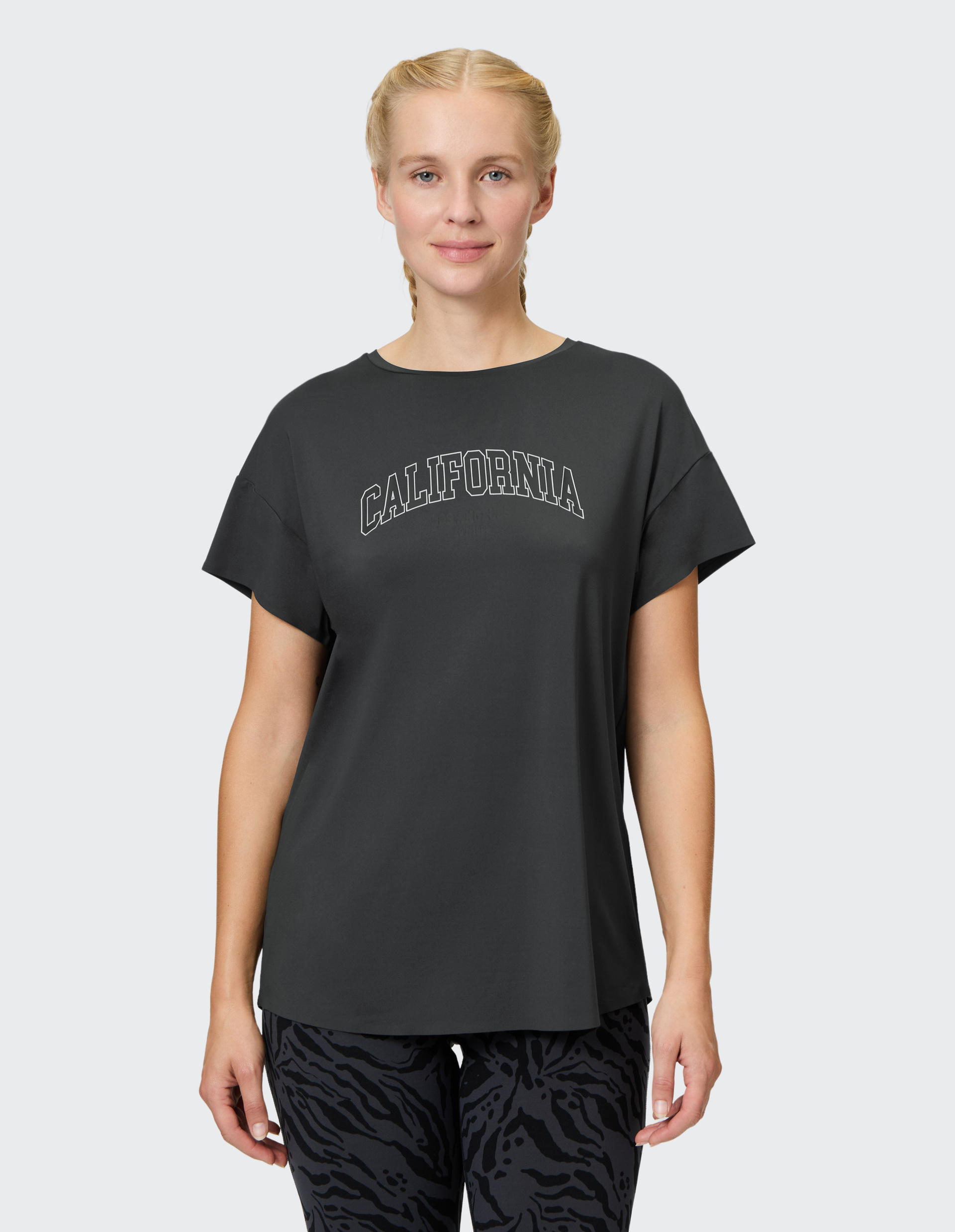 Eine Person trägt ein dunkelgraues T-Shirt mit dem Aufdruck "California" und eine gemusterte dunkle Hose, posiert vor neutralem Hintergrund.