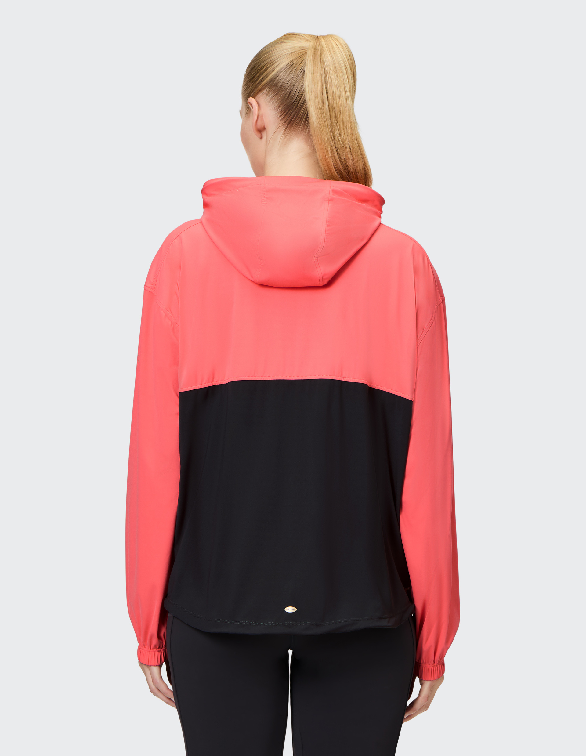 Frau mit Pferdeschwanz trägt eine zweifarbige Jacke in Pink und Schwarz mit Kapuze, von hinten gesehen, vor hellem Hintergrund.