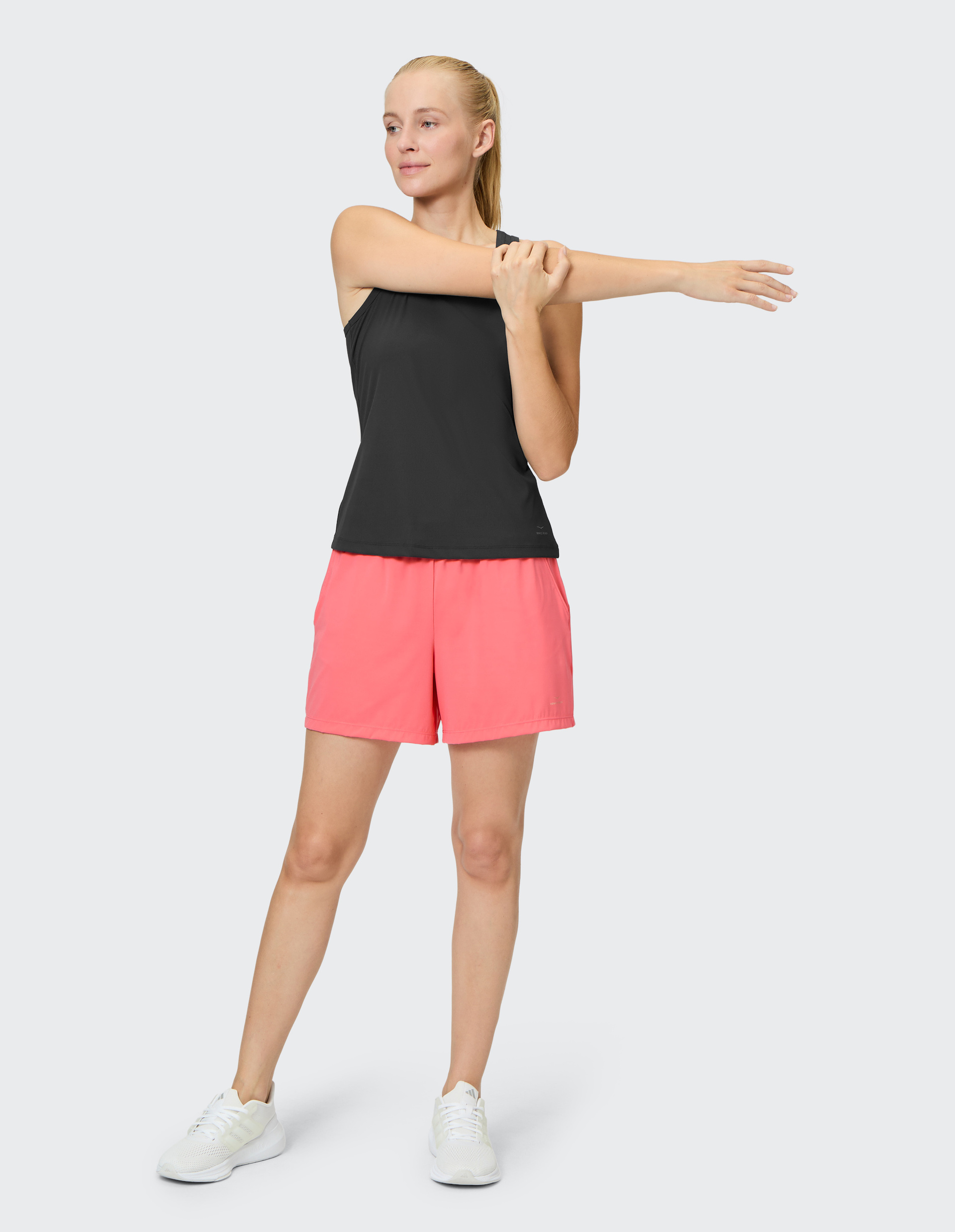 Frau in schwarzem Top und pinken Shorts dehnt im Stehen den rechten Arm über der Brust, vor grauem Hintergrund.