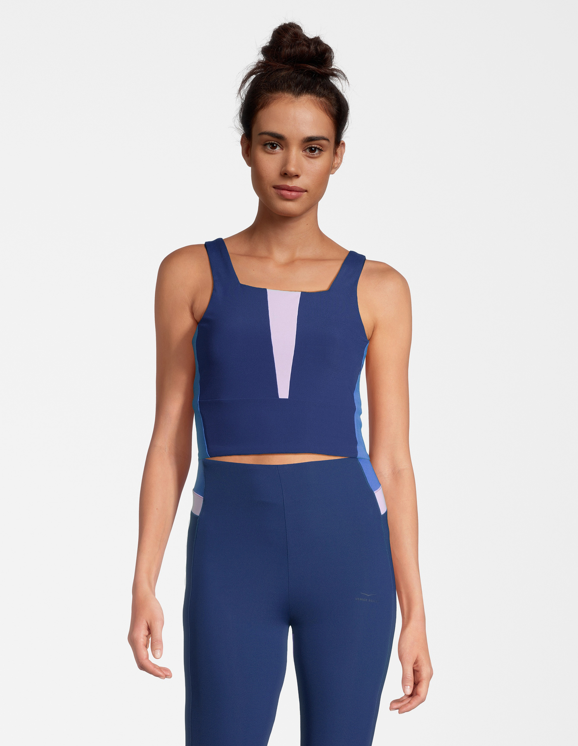 Frau in blau-lila Sportkleidung mit Träger-Top und langer Hose vor weißem Hintergrund, Haare in einem Dutt.