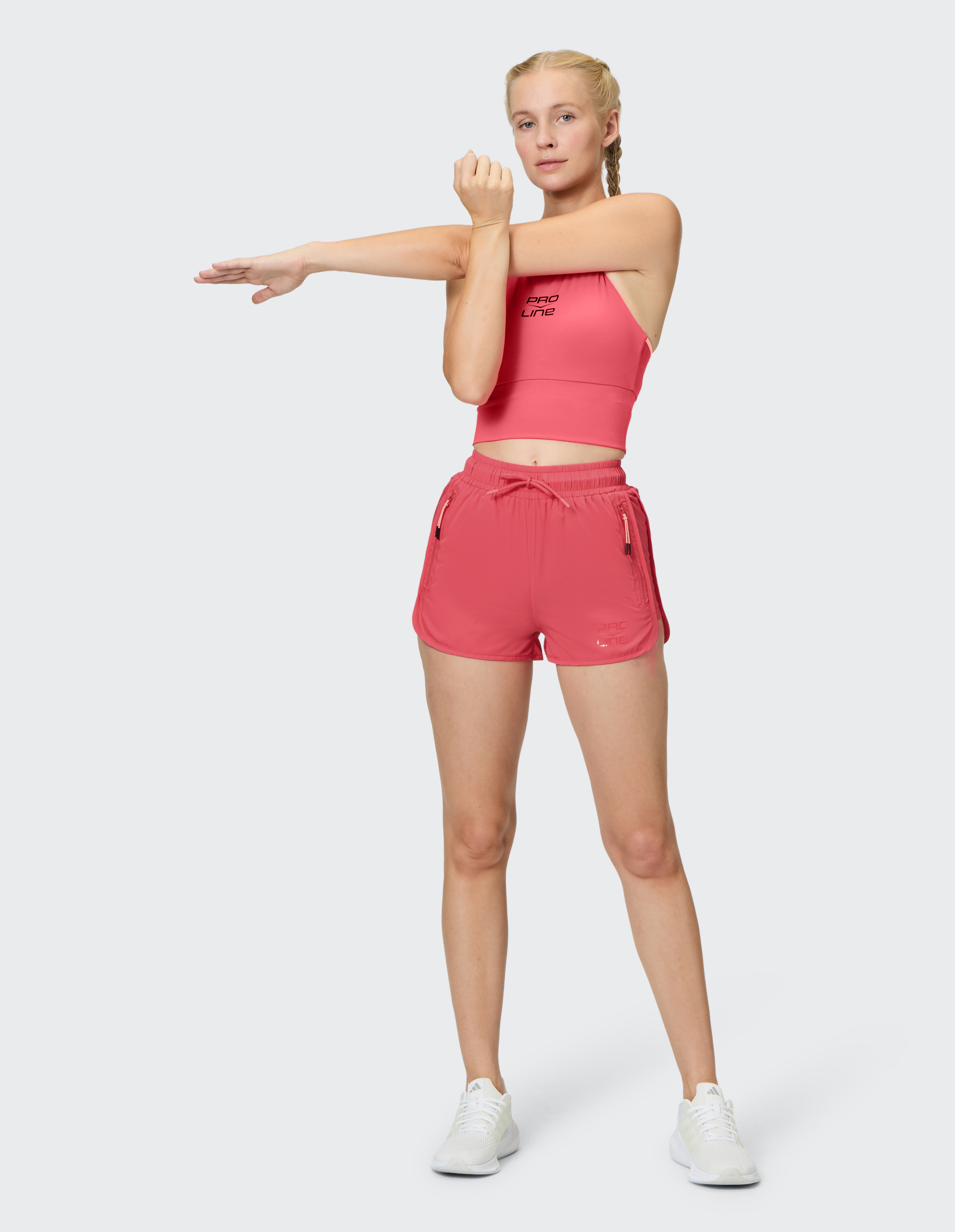 Eine Frau dehnt ihre Arme, trägt rosa Sportkleidung und weiße Turnschuhe vor einem neutralen Hintergrund.