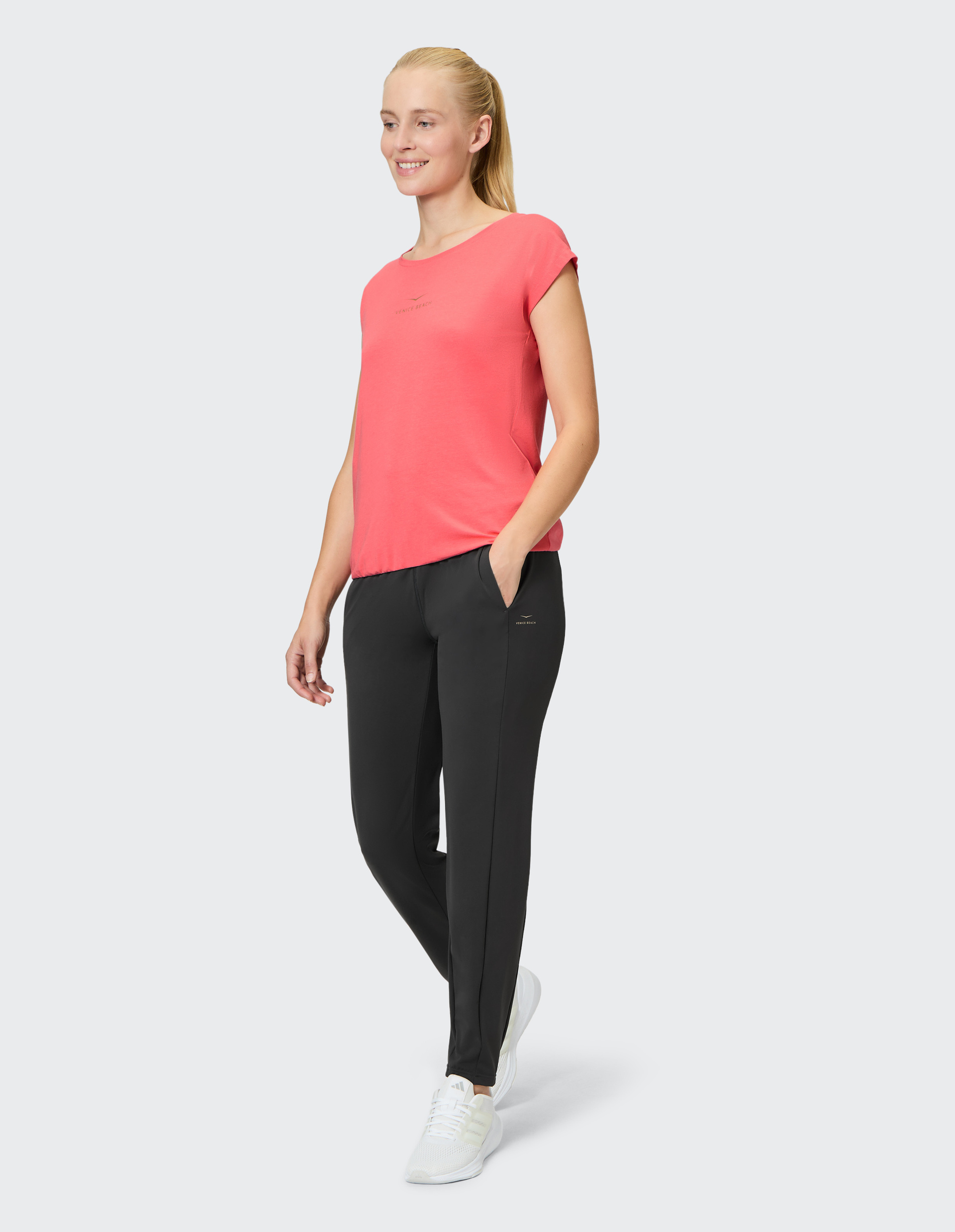 Frau in pinkem T-Shirt und schwarzen Hosen, eine Hand in der Tasche, lächelnd, trägt weiße Sneaker, steht vor hellem Hintergrund.